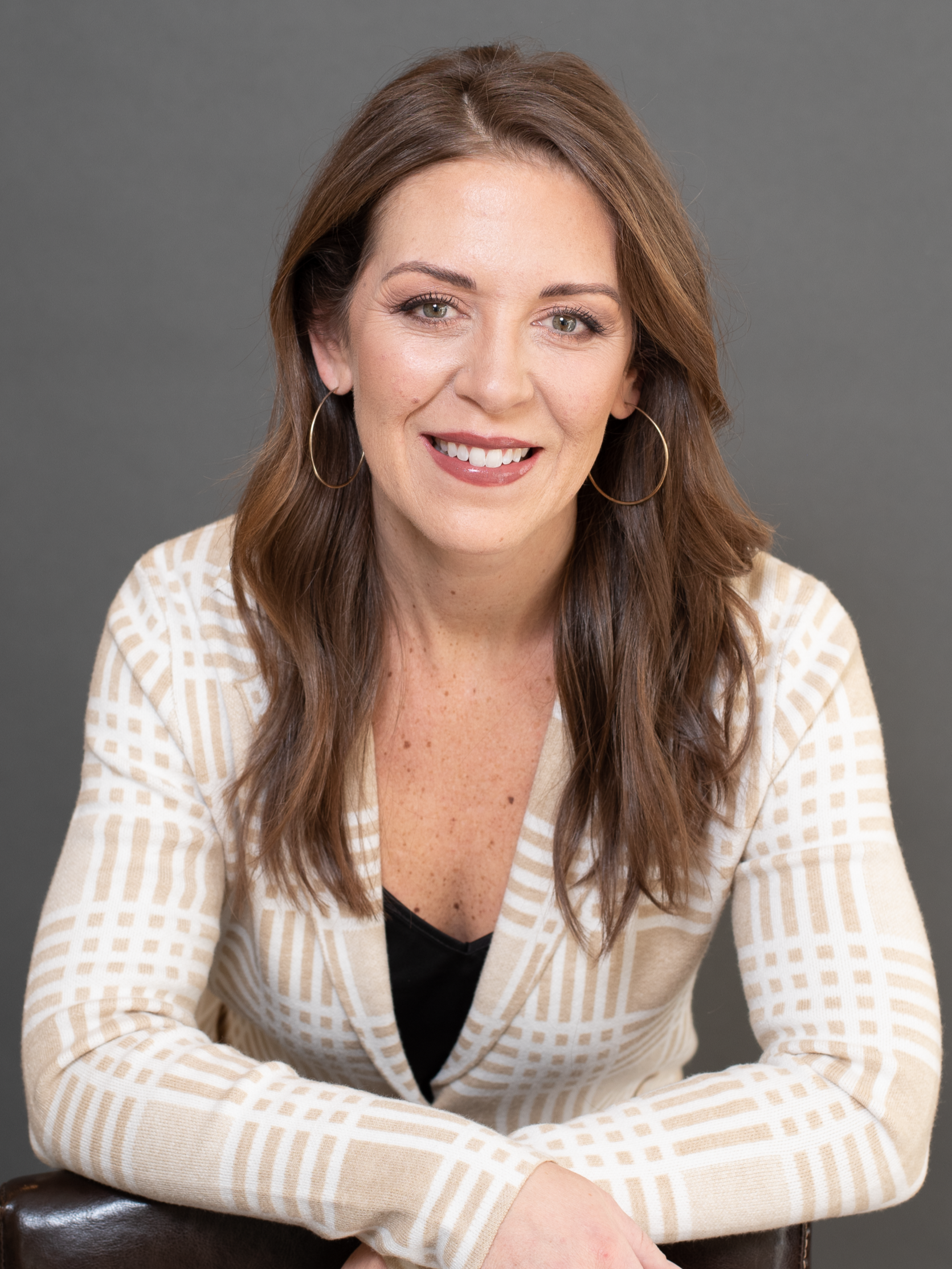A woman with long brown hair, wearing hoop earrings and a beige and white checkered blazer, is sitting behind a dark brown chair, smiling, against a plain gray background.