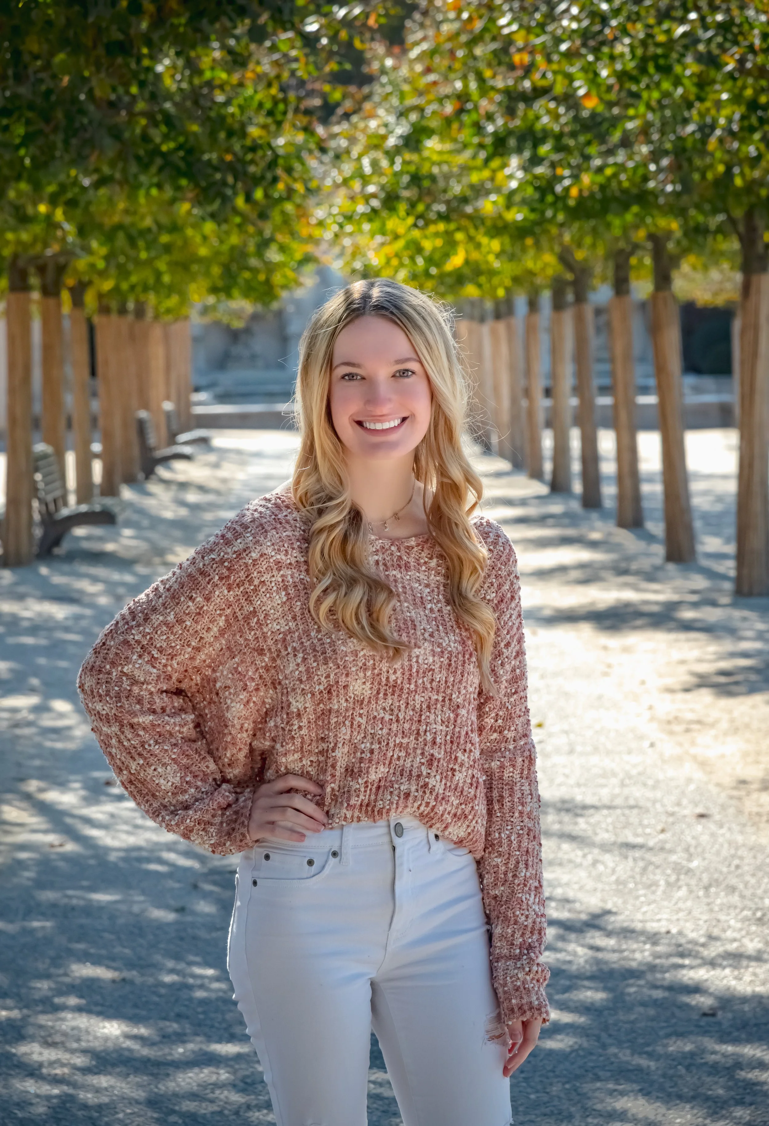 A young woman with blonde curly hair and a bright smile stands outdoors on a sunny day, wearing a pink knit sweater and white pants. She is positioned on a pathway lined with wooden posts and benches, with lush green trees in the background.