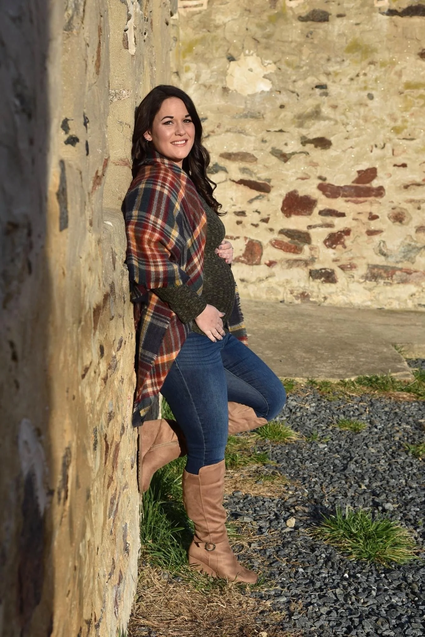 A woman leaning against a stone wall outdoors, wearing a plaid shawl, dark top, blue jeans, and tan boots, with greenery and gravel on the ground.