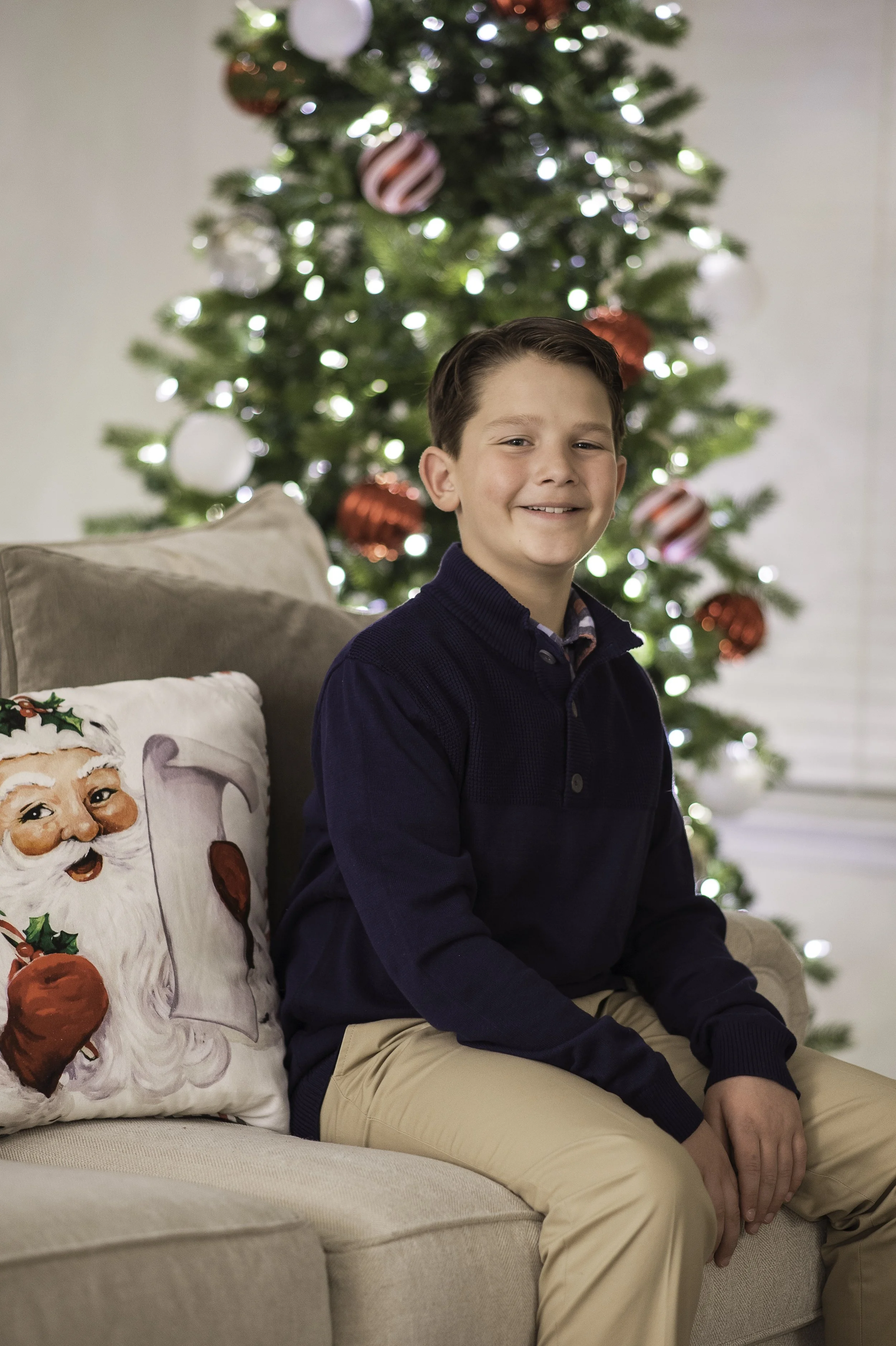 A young boy with brown hair, wearing a navy blue sweater and khaki pants, is sitting on a beige couch with a decorative Christmas pillow featuring Santa Claus and holly. Behind him, a decorated Christmas tree with white and red ornaments and lights i