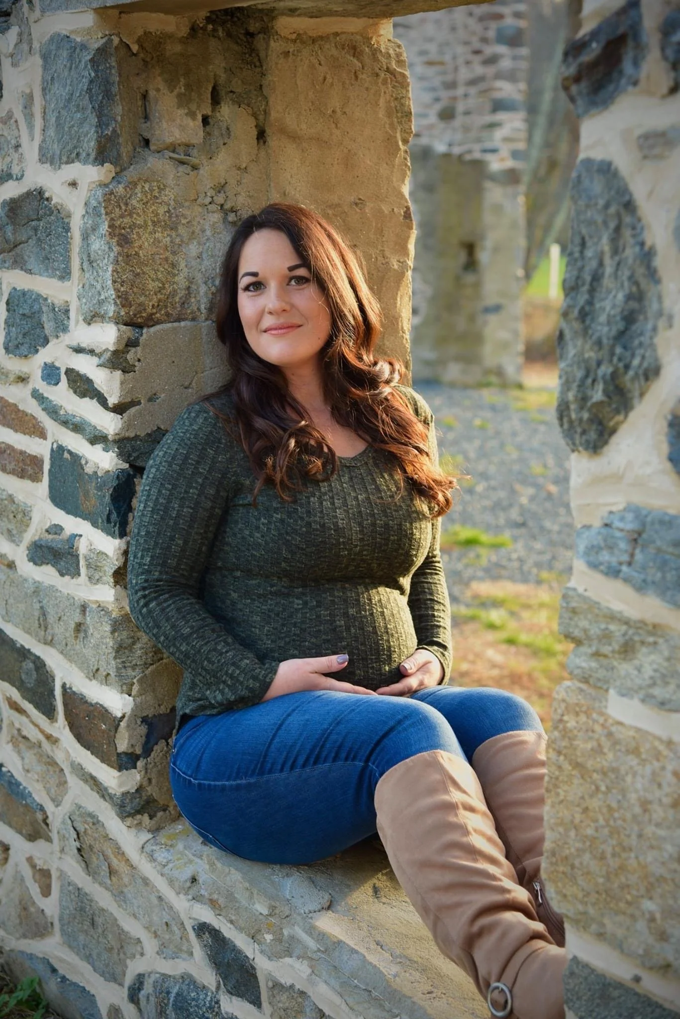 A woman with long dark hair, sitting on a stone wall, looking at the camera, wearing a green patterned sweater, blue jeans, and beige boots, with a natural outdoor background.