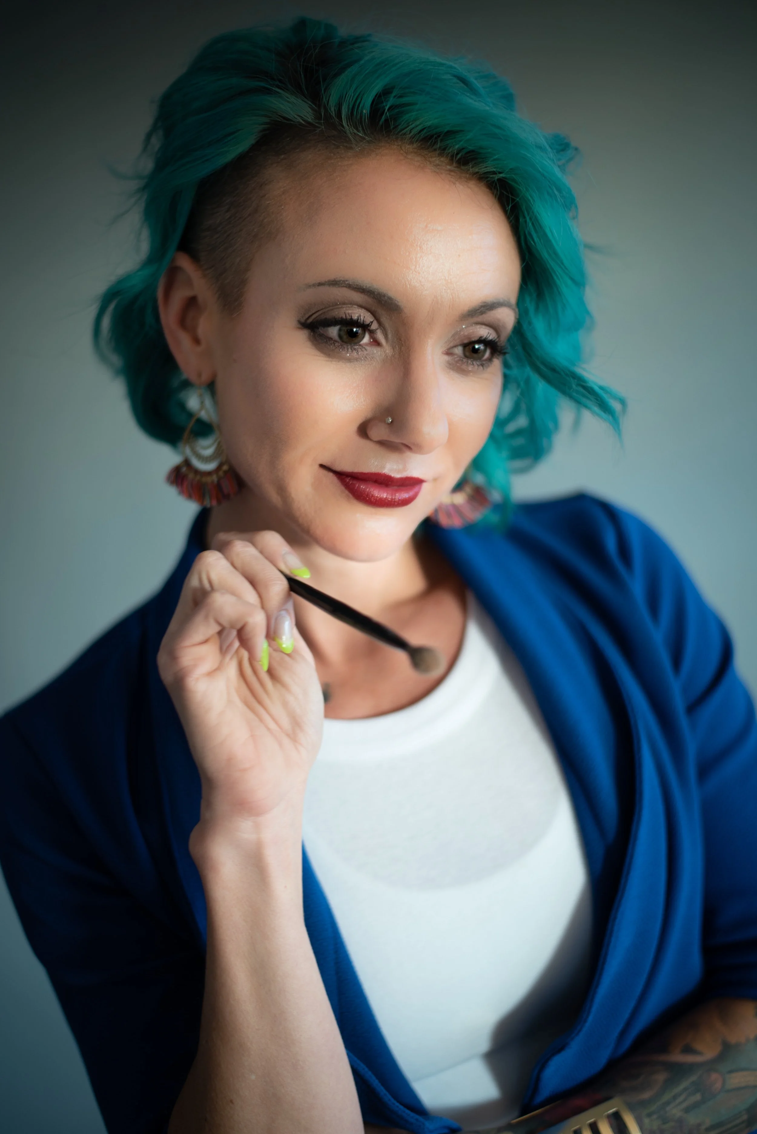 A woman with blue-green hair and tattoos is looking down, holding a makeup brush near her face, wearing a white shirt and a blue jacket.