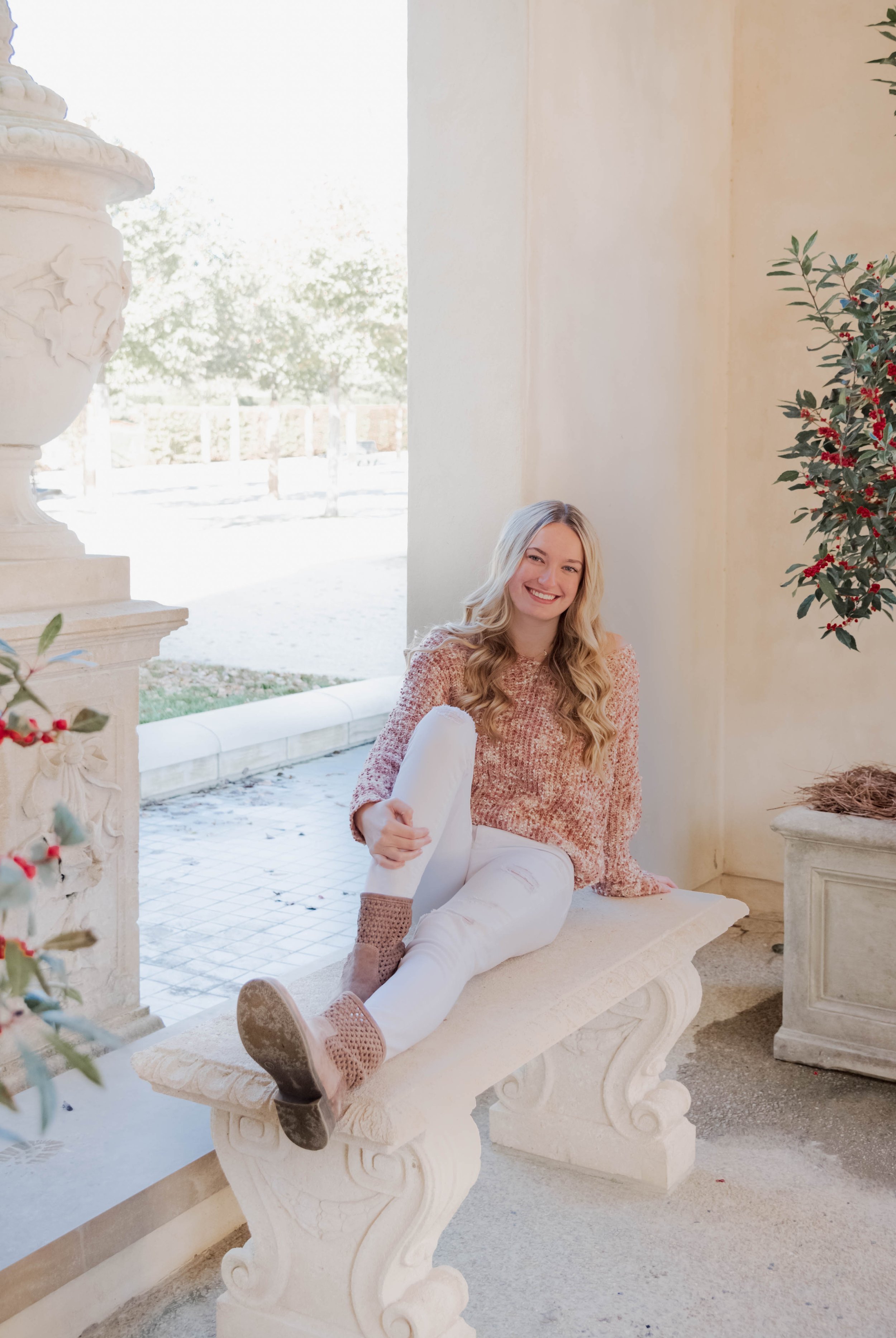 A young woman with long blonde hair sitting on a decorative stone bench, smiling, wearing a pink sweater, white ripped jeans, and tan boots, in an outdoor setting with a large urn, greenery, and a tree in the background.