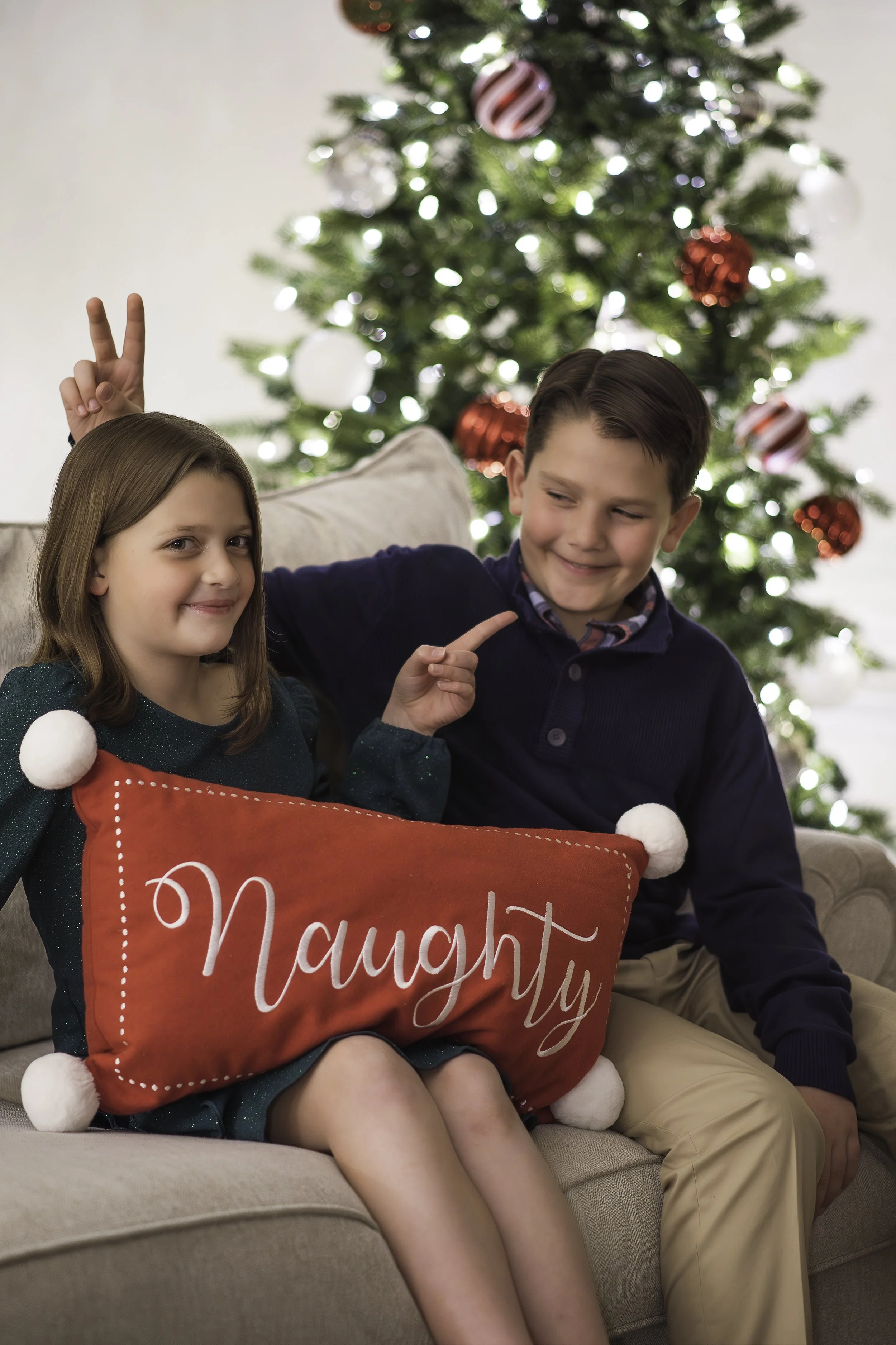 Two children sitting on a beige couch in front of a decorated Christmas tree. The girl, wearing a green dress with white pom-pom details, is holding a red pillow with the word 'Naughty' and white pom-poms at the corners. The boy, wearing a navy sweat