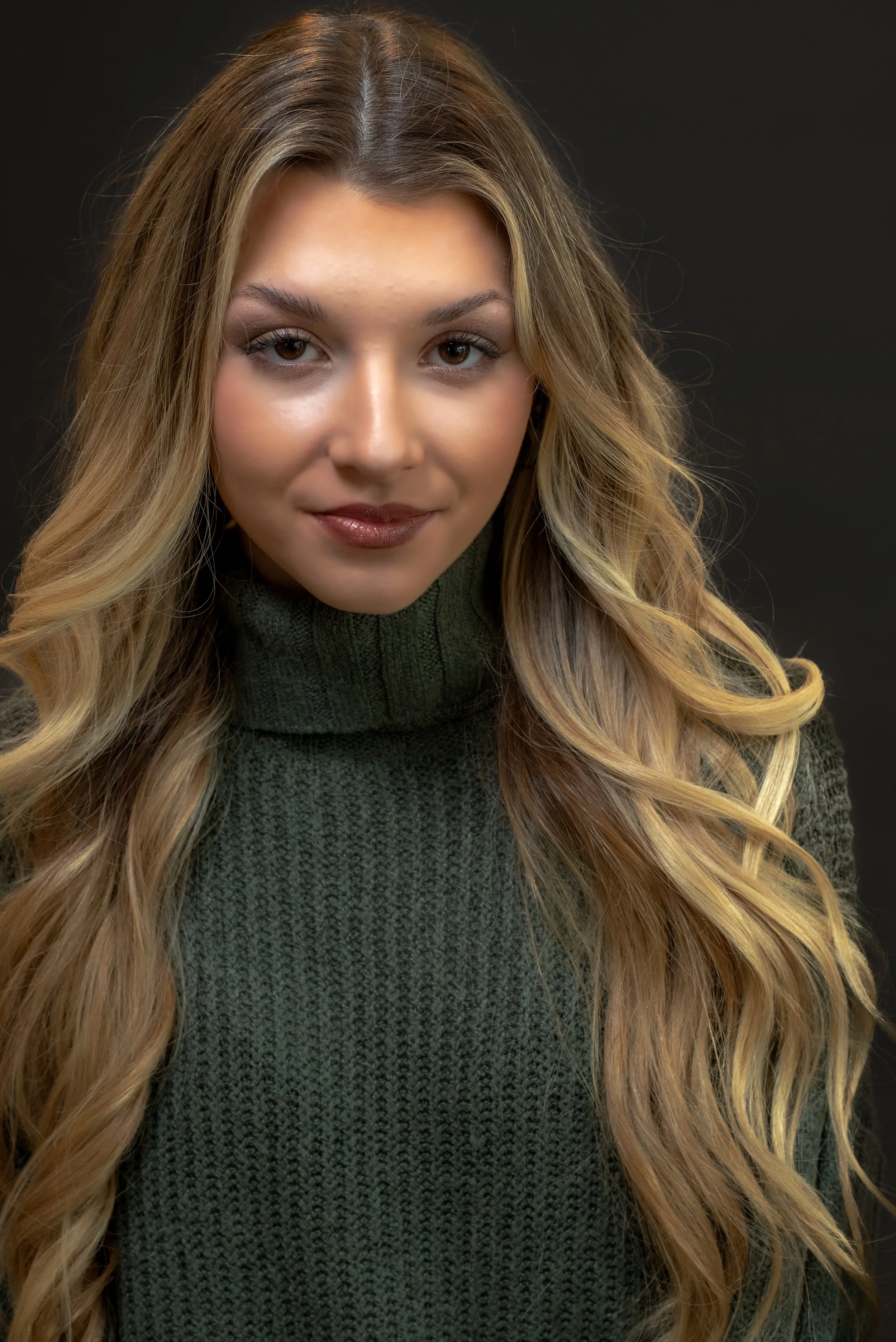 A woman with long, wavy blonde hair wearing a dark green sweater, looking directly at the camera with a slight smile, against a dark background.