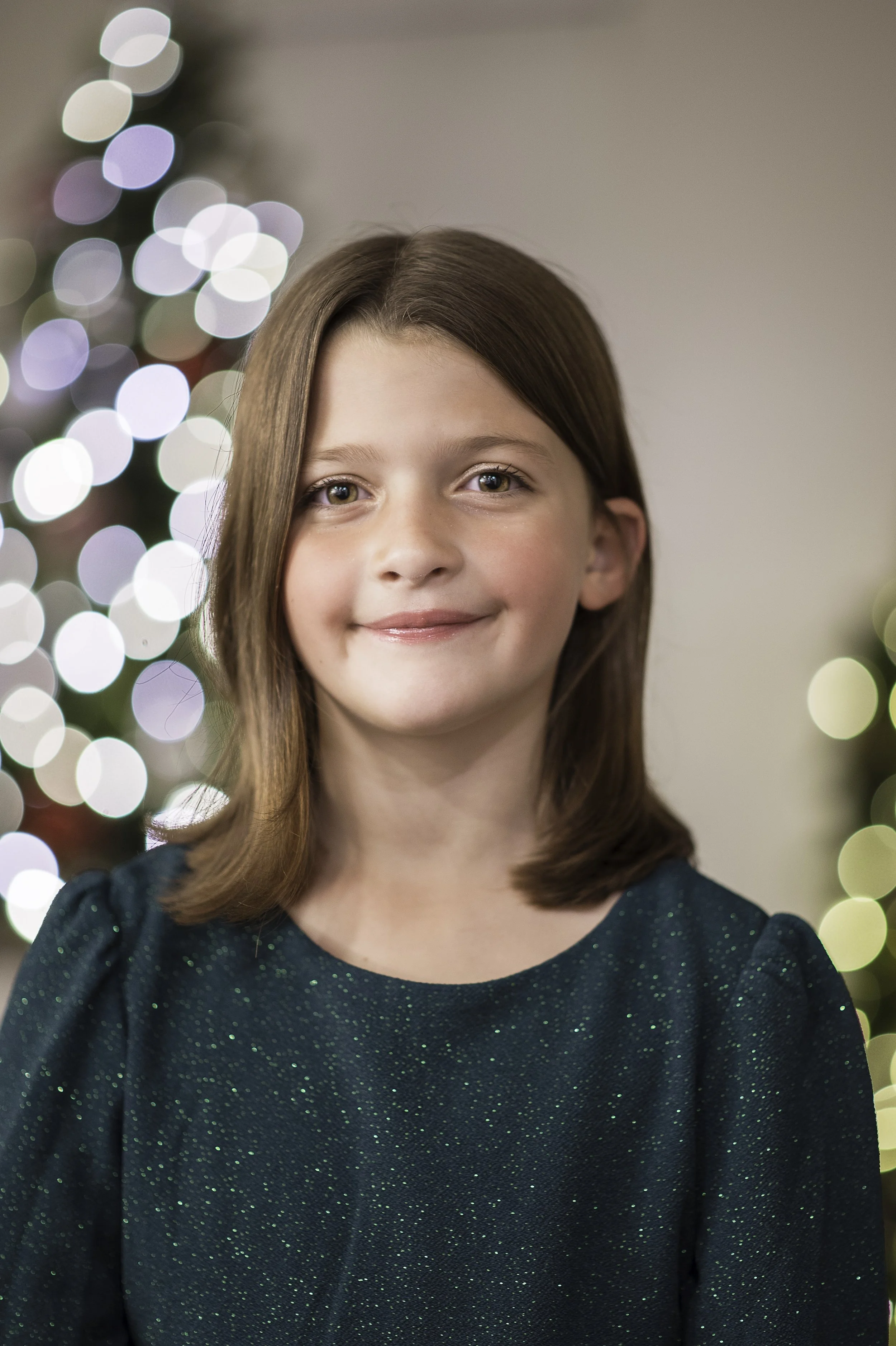 A young girl with brown hair and hazel eyes wearing a dark green, sparkly dress, smiling at the camera. Out-of-focus lights in the background suggest a festive holiday setting.
