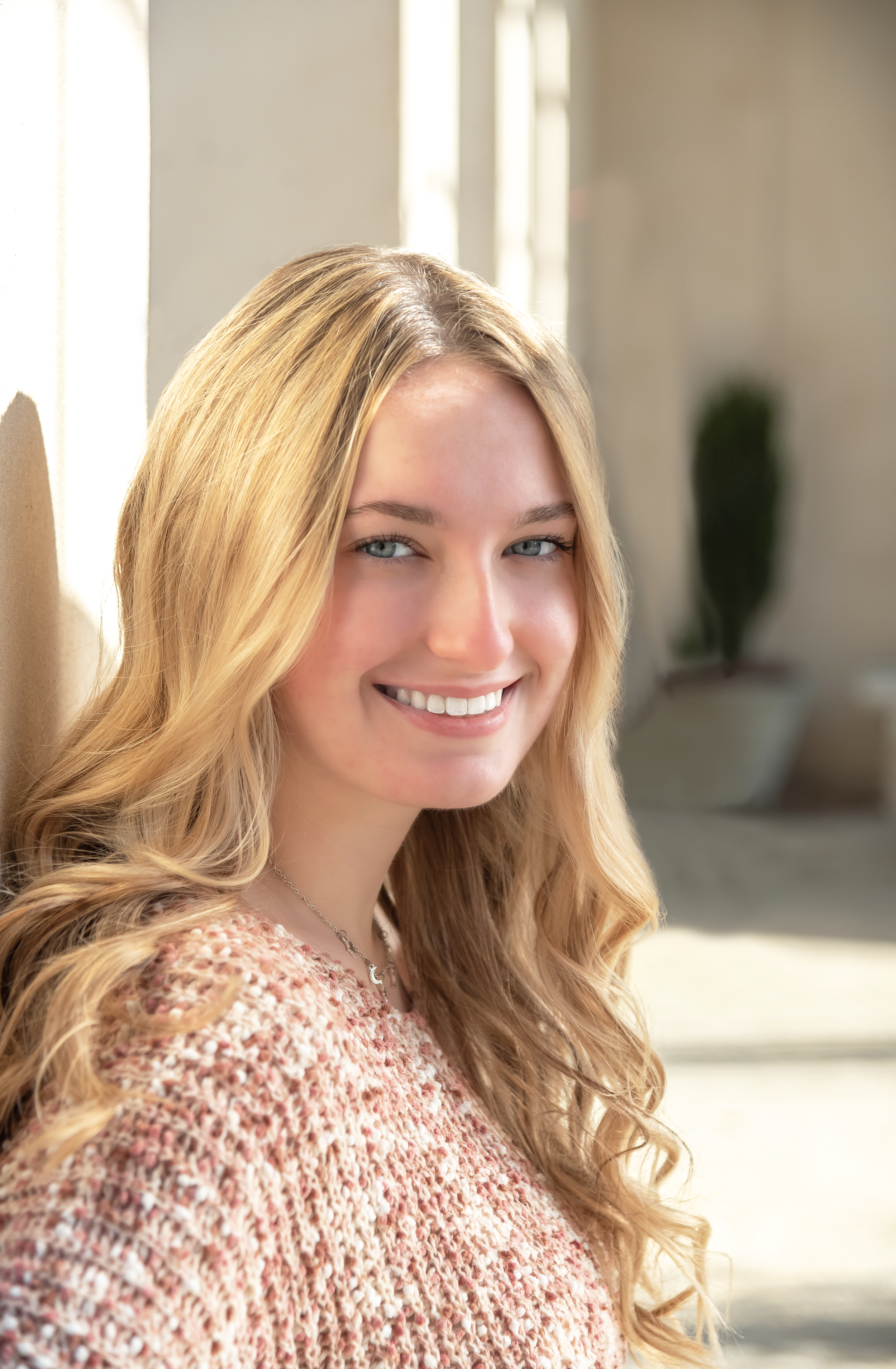 Close-up of a smiling young woman with long wavy blonde hair, blue eyes, wearing a pink textured sweater, standing indoors near a window with natural light, with a blurred green plant in the background.