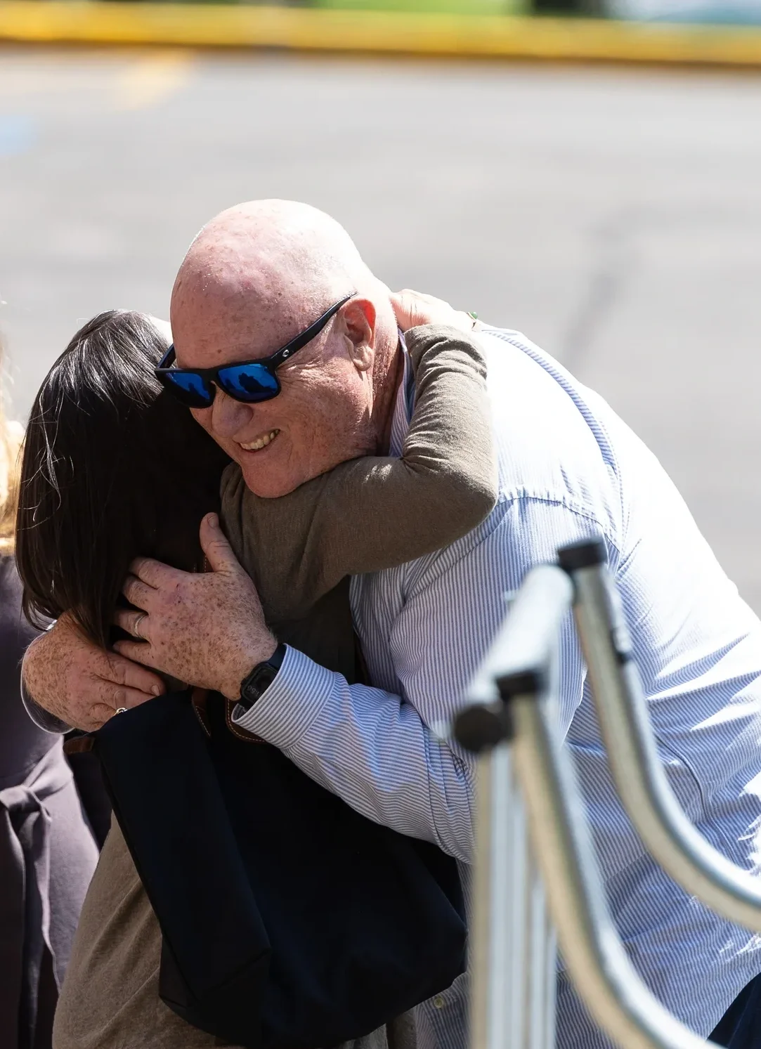 A white balding middle-aged man wearing sunglasses (Elliott Haverlack) hugs a woman with dark hair and a brown sweater. The photo is taken at close range and we cannot see the woman's face or features.