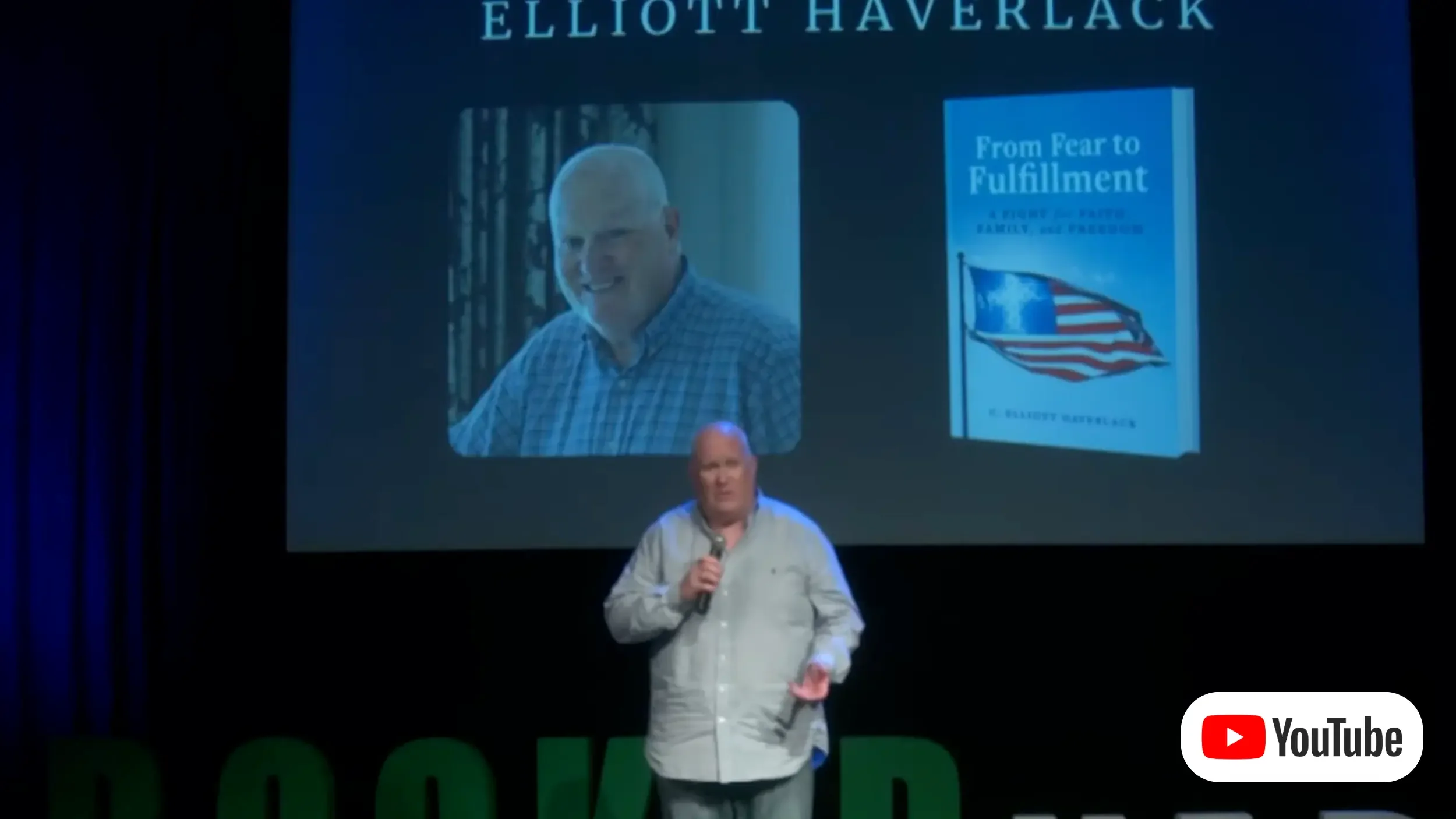 A man in a blue striped button down shirt and jeans holds a microphone in his right hand as he stands onstage and speaks. The projector screen behind him shows a photo of him alongside his book From Fear to Fulfillment and his name Elliott Haverlack.