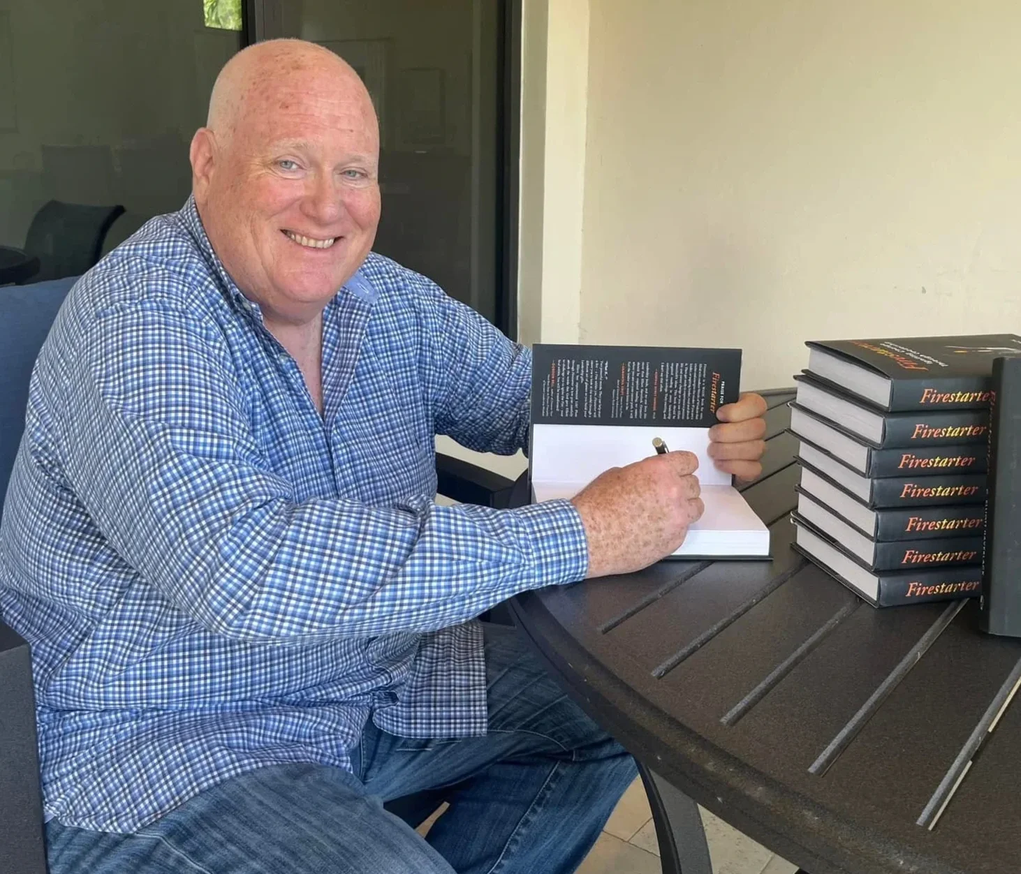 A middle-aged bald white man in a blue plaid shirt sits at an outdoor patio table signing a copy of Firestarter. A stack of Firestarter books sits on the table in front of him.