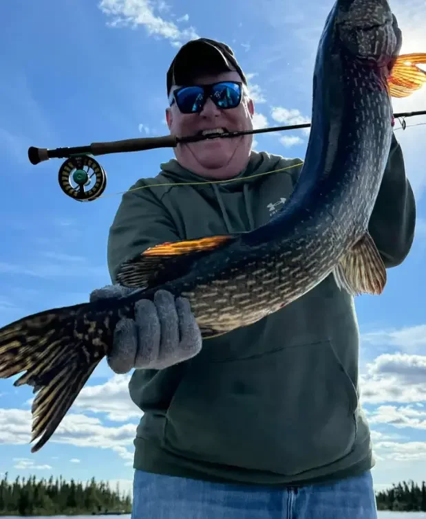 Photo of a middle-aged white man wearing a dark sage green hoodie, sunglasses, a hat, and jeans with a fly fishing rod held in his teeth holding up a fish