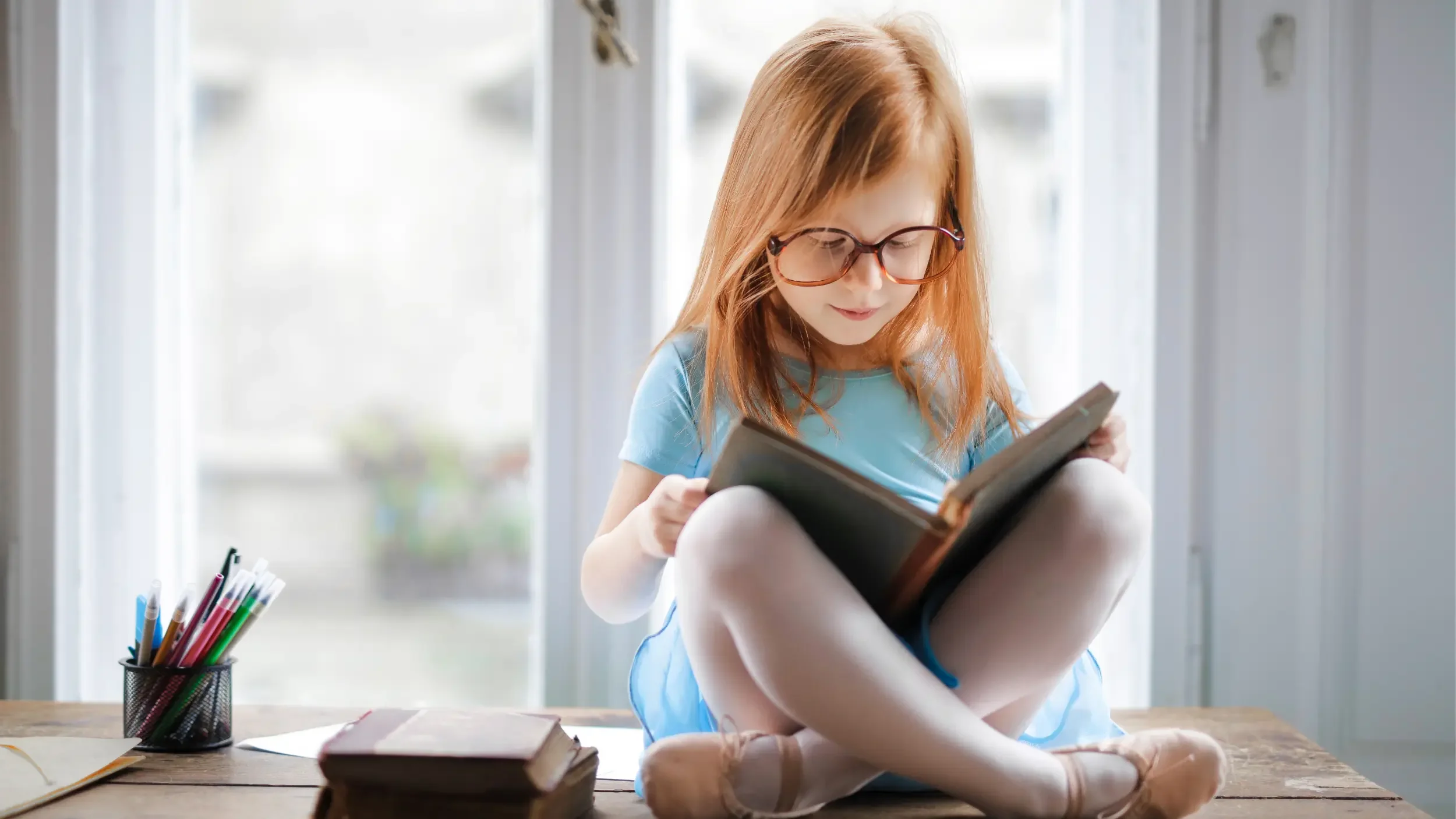 A small caucasian girl with red hair and large brown glasses, wearing a blue ballet dress, tights, and ballet slippers sits cross-legged on a desk reading a book