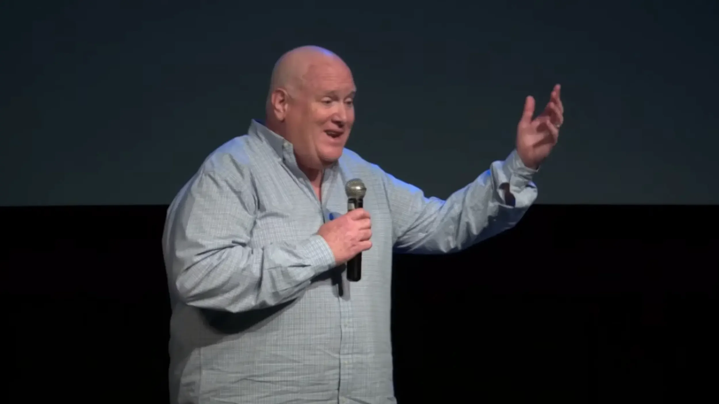 Close-up of Elliott Haverlack speaking - a bald caucasian male with a long sleeved light blue button down holding a microphone and gesturing broadly.