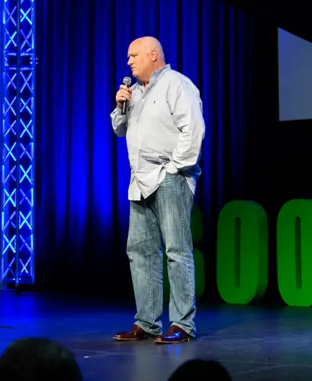 C. Elliott Haverlack at Booked Naples 2022. A caucasian man in jeans and a long sleeve button down holds a microphone and speaks onstage.