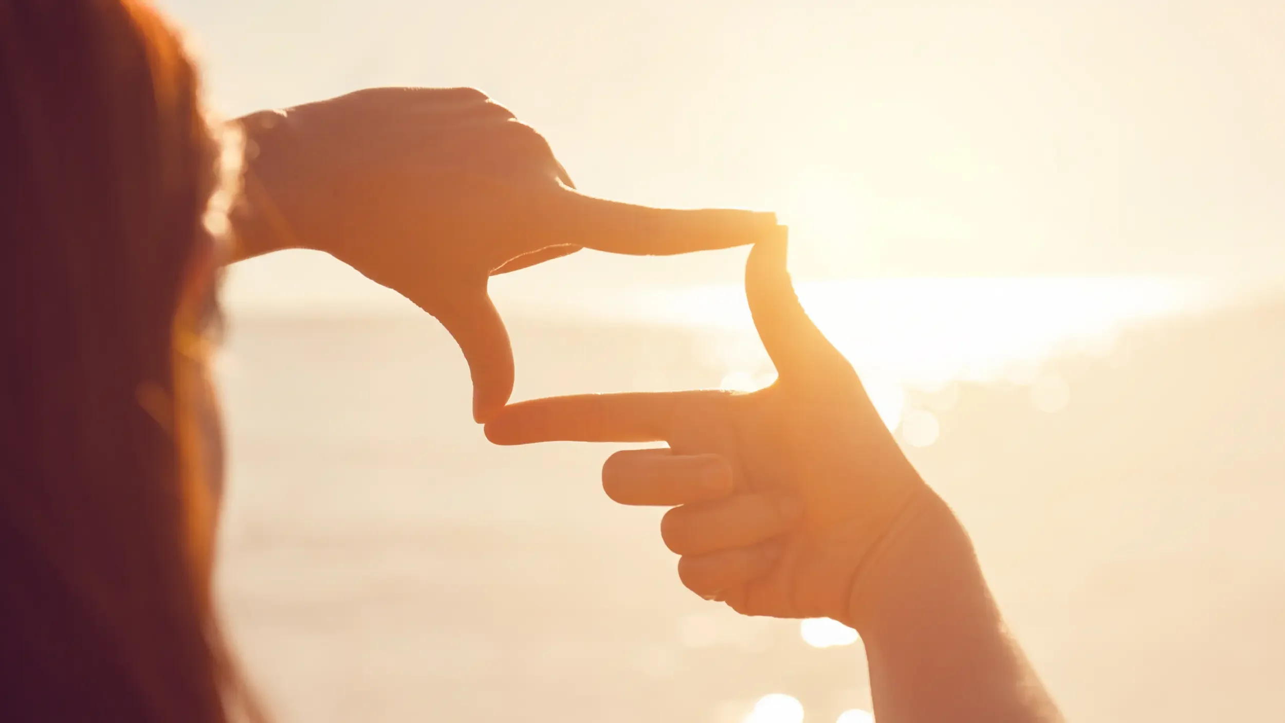 close-up from behind of a woman with her fingers making a frame looking over a body of water towards a sunset