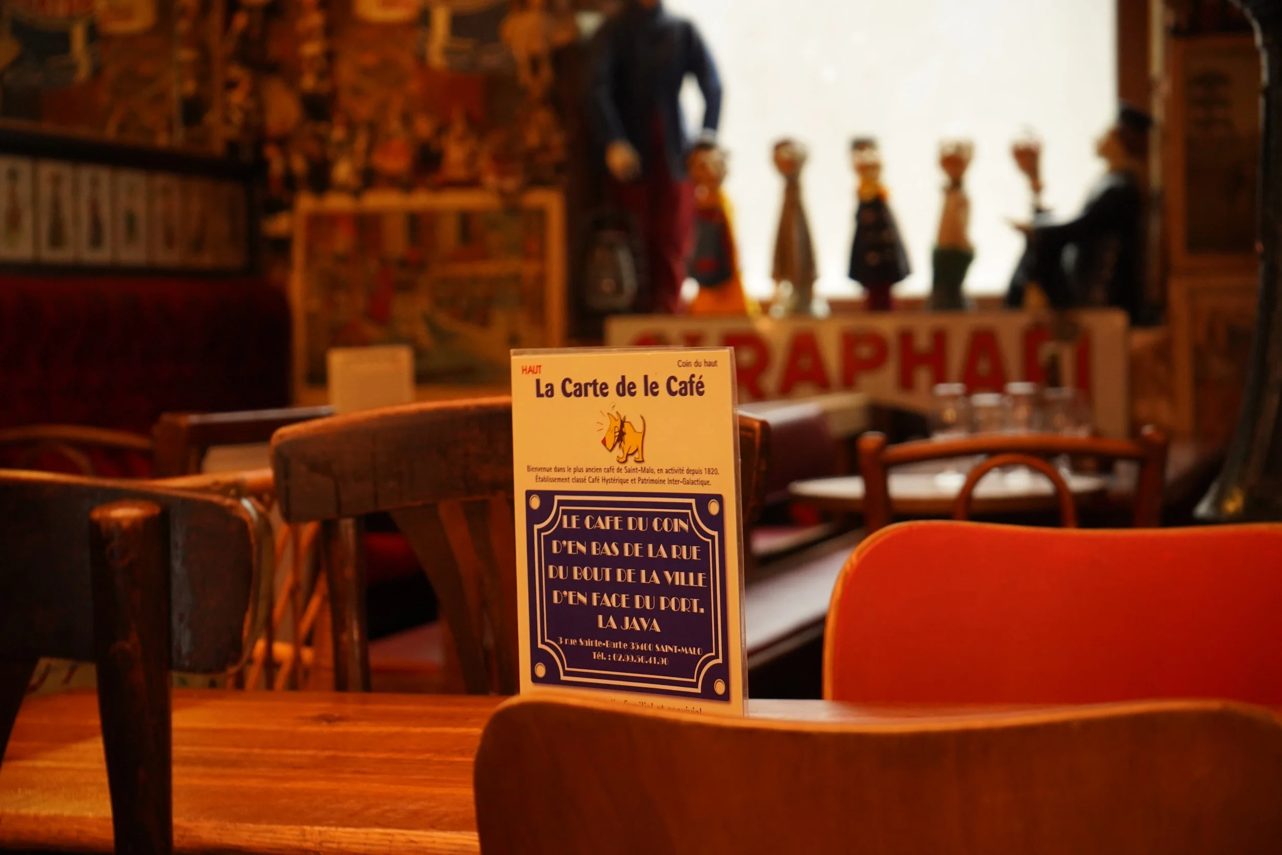 Une table en bois dans un café avec un menu en français placé dessus. En arrière-plan, il y a des dessins de mannequin et une personne assise en train de lire ou écrire.