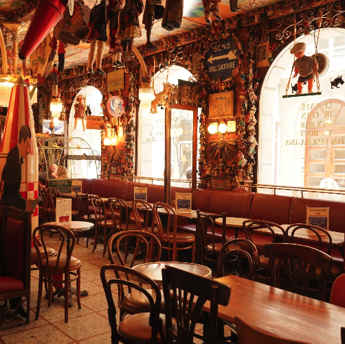 Intérieur d'un café ou restaurant coloré avec des chaises en bois, des tables, des murs décorés avec des objets suspendus et des lumières chaleureuses, fenêtres lumineuses laissant entrer la lumière naturelle.