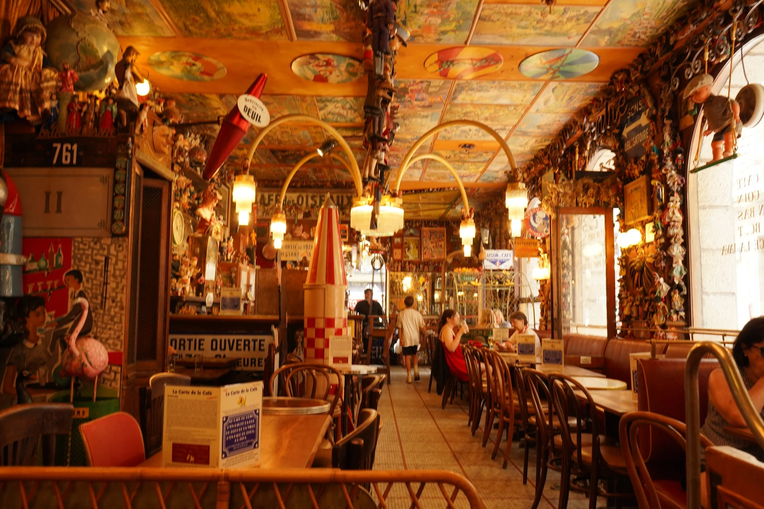 Intérieur d'un restaurant ou café avec décor coloré, figurines et affiches sur les murs, tables en bois avec menus, plusieurs personnes assises.
