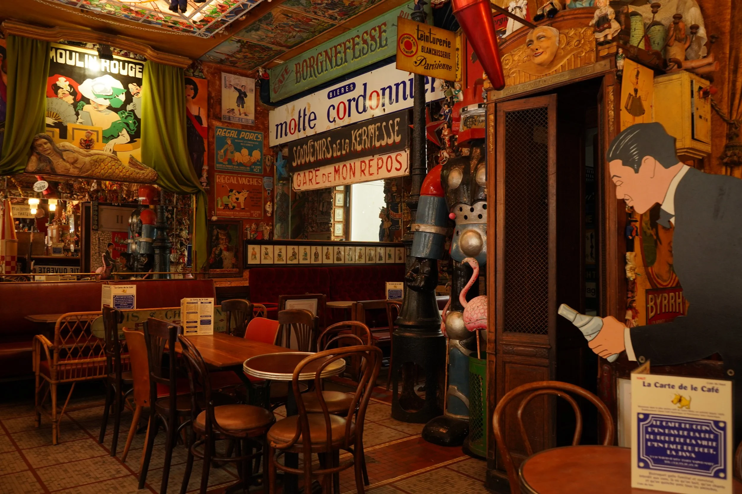 Intérieur d'un café ou bar/pub avec décor vintage et comics, affiches colorées, statues et objets d'époque, tables et chaises en bois.