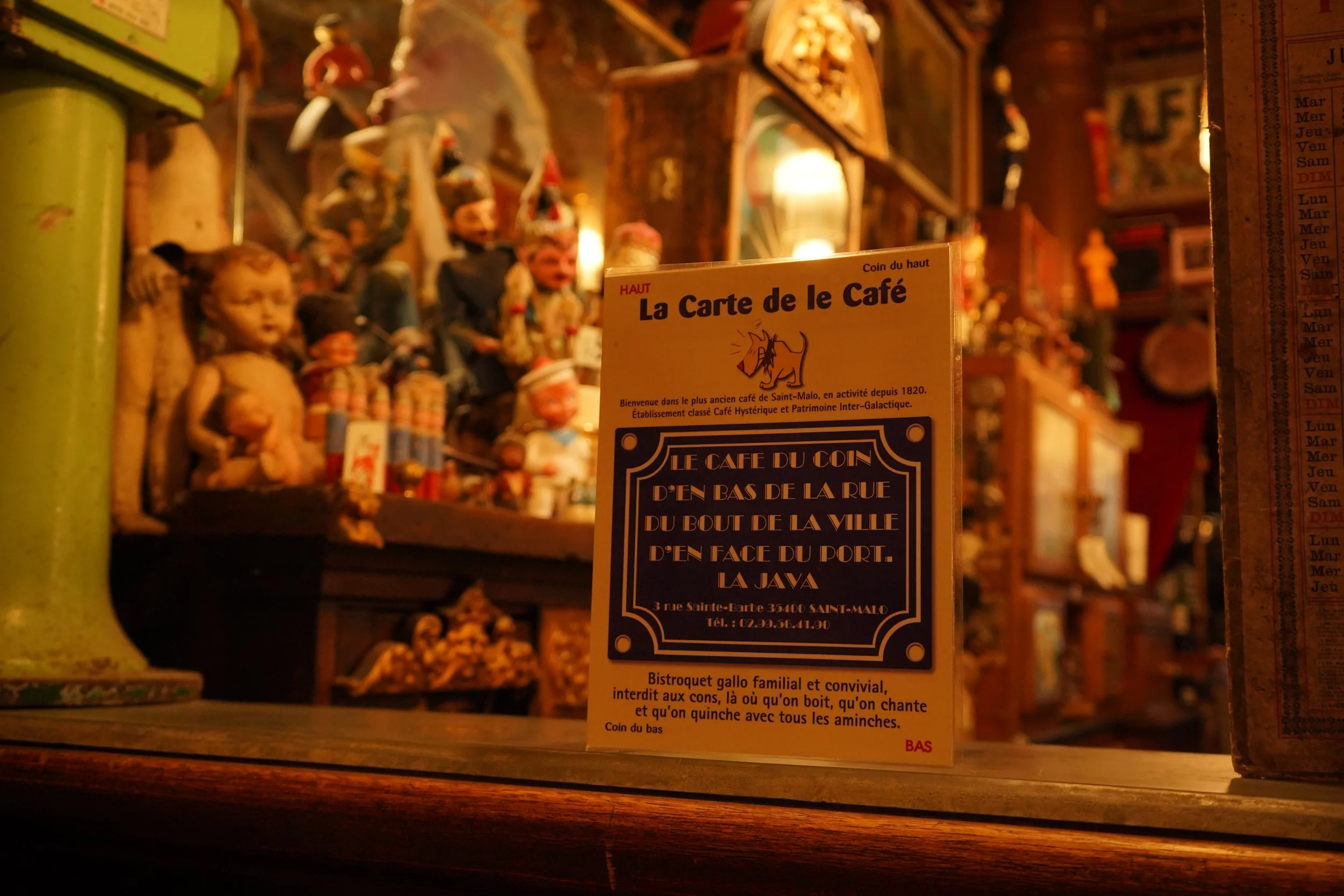 Carte des boissons dans le café du coin posé sur le comptoir du barre avec déco brocante ave objets et poupées derrière