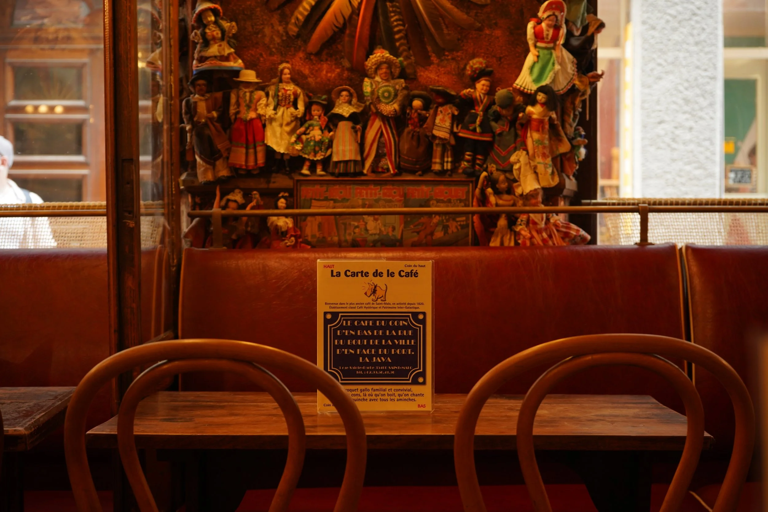 Une décoration murale traditionnelle avec des figurines en tissu représentant des personnages en costumes traditionnels dans un cadre en bois. La scène est située derrière une table en bois avec des chaises en bois en premier plan. Un panneau d'information en français est placé sur la table.