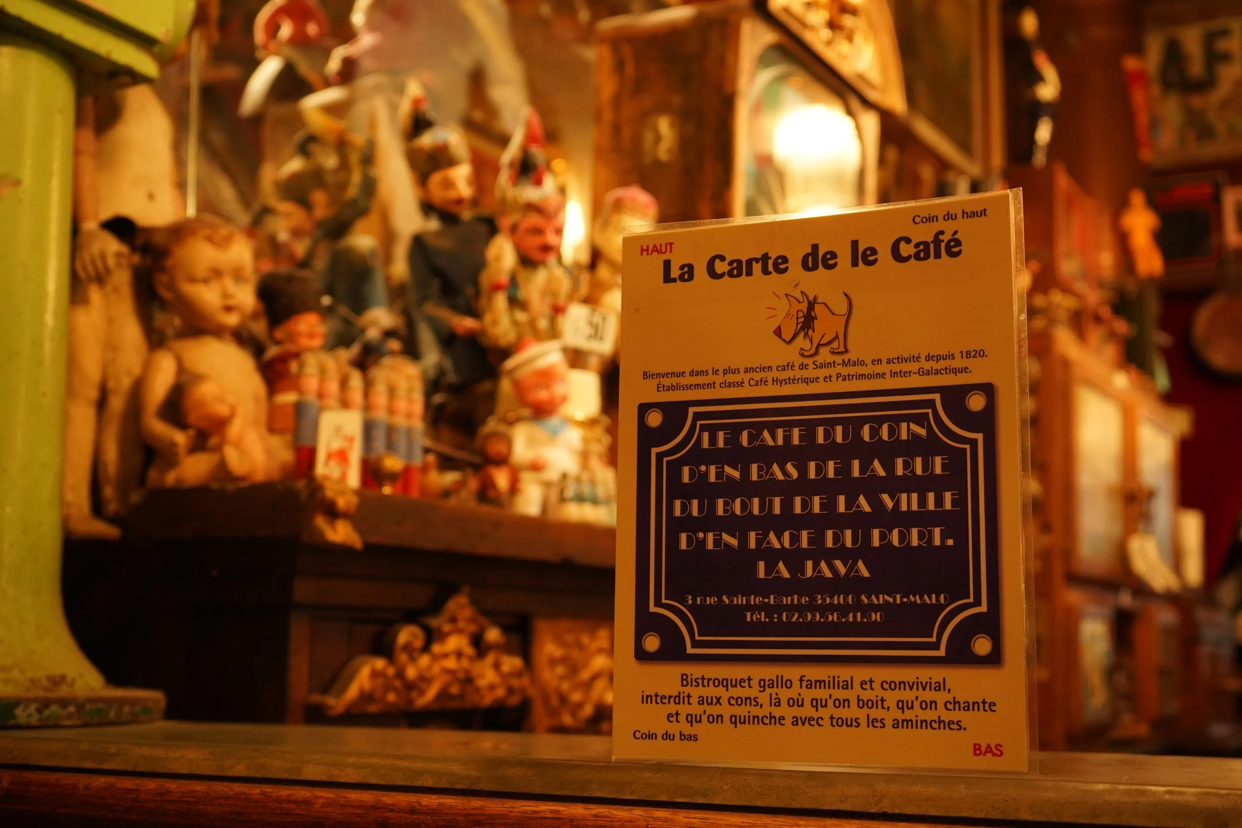 Une affiche présente dans un café ancien, avec des figurines en arrière-plan, dont des marionnettes ou des poupées. La affiche mentionne La Carte du Café et fournit des détails sur l'établissement, situé à Sainte-Malo, avec un décor riche et vintage.