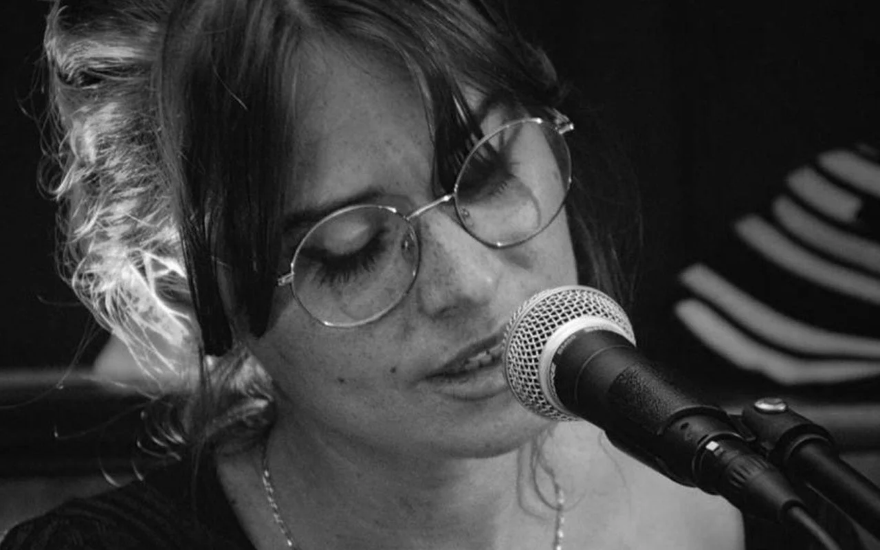 Une femme avec des lunettes rondes, des cheveux courts, chante ou parle dans un microphone, en noir et blanc.