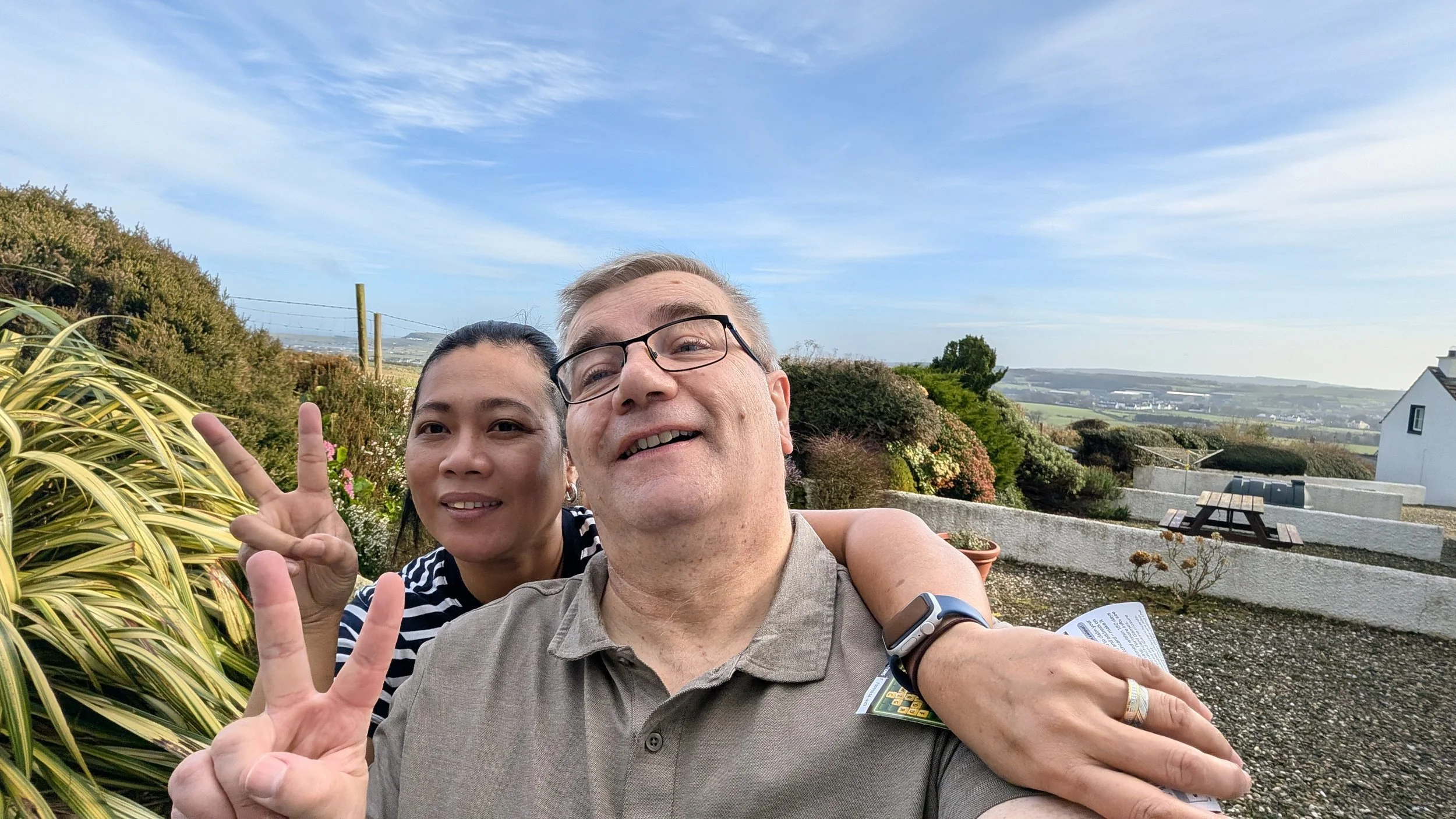 Two people taking a selfie outdoors, with a woman making a peace sign and smiling, and a man standing close, both smiling, in a garden or patio setting with trees, bushes, and a blue sky in the background.