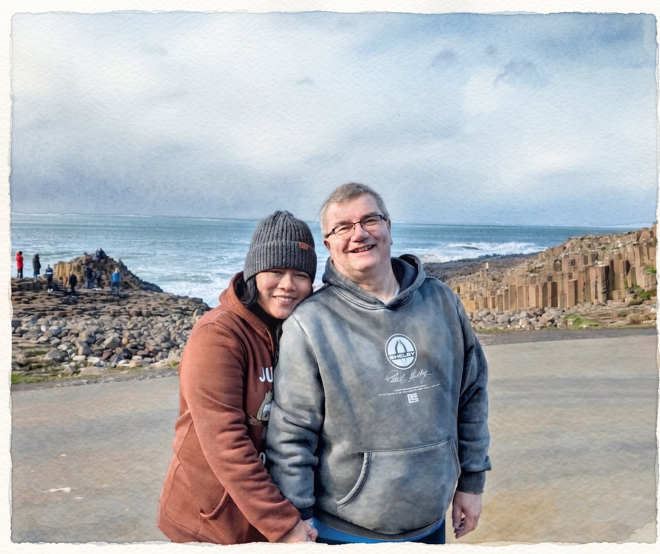 Two people standing on a rocky seashore, smiling at the camera, with a man wearing glasses and a gray hoodie, and a woman wearing a brown jacket and gray beanie, with a cliffside in the background and an overcast sky.