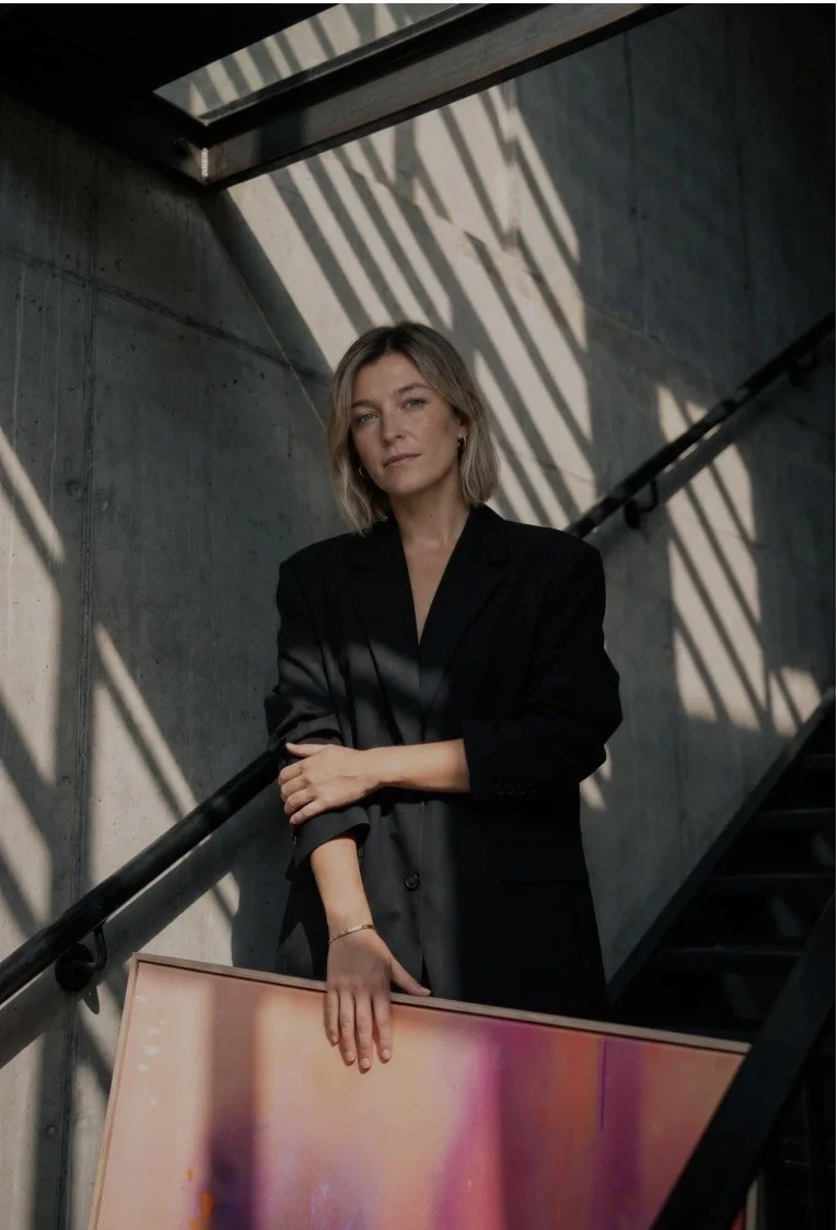 Eine Frau steht auf einer Treppe in einem Raum mit rohem Betonwänden, während Sonnenlicht Schatten an die Wand wirft, sie trägt einen schwarzen Anzug.