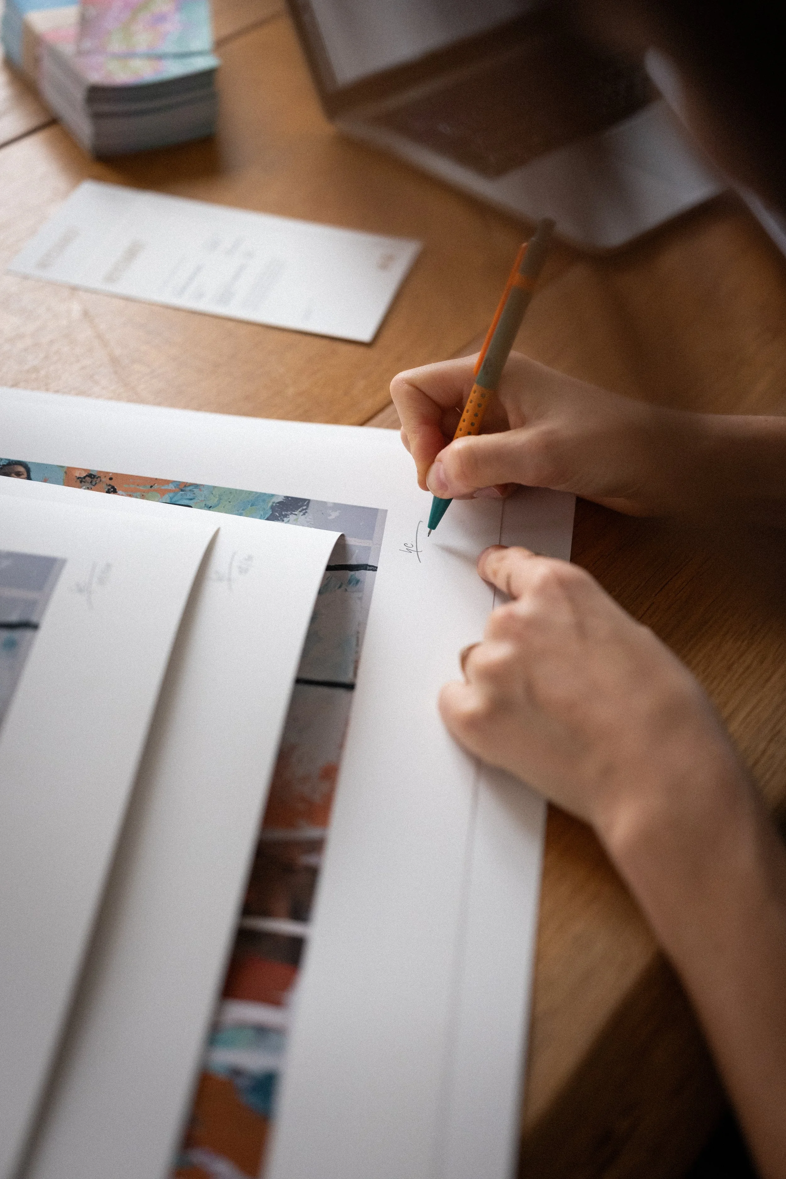 Eine Hand unterschreibt einen Fine Art Print, dahinter sind weitere zu sehen. Im Hintergrund liegen gedruckte Dokumente und ein Apple Laptop auf einem Holztisch.