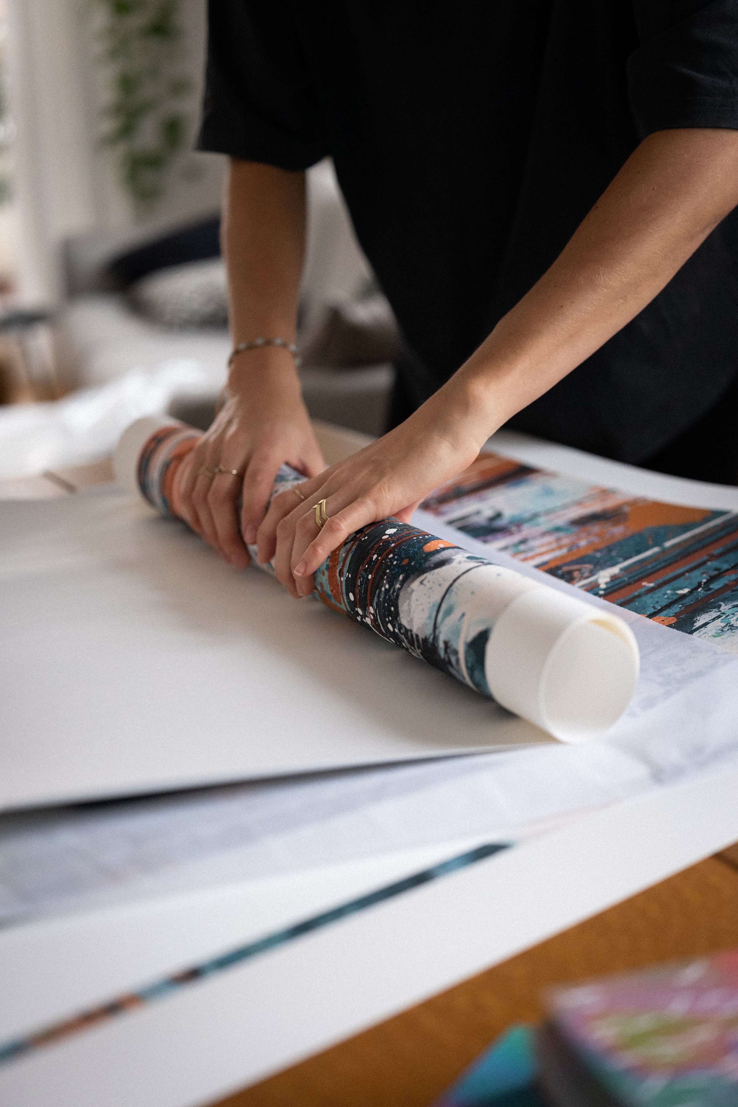 Person rollt ein gerolltes Poster auf einem weißen Tisch. Die Person trägt eine schwarze Kleidung und trägt mehrere Ringe und eine Armband. Im Hintergrund sind Möbel und Pflanzen sichtbar.