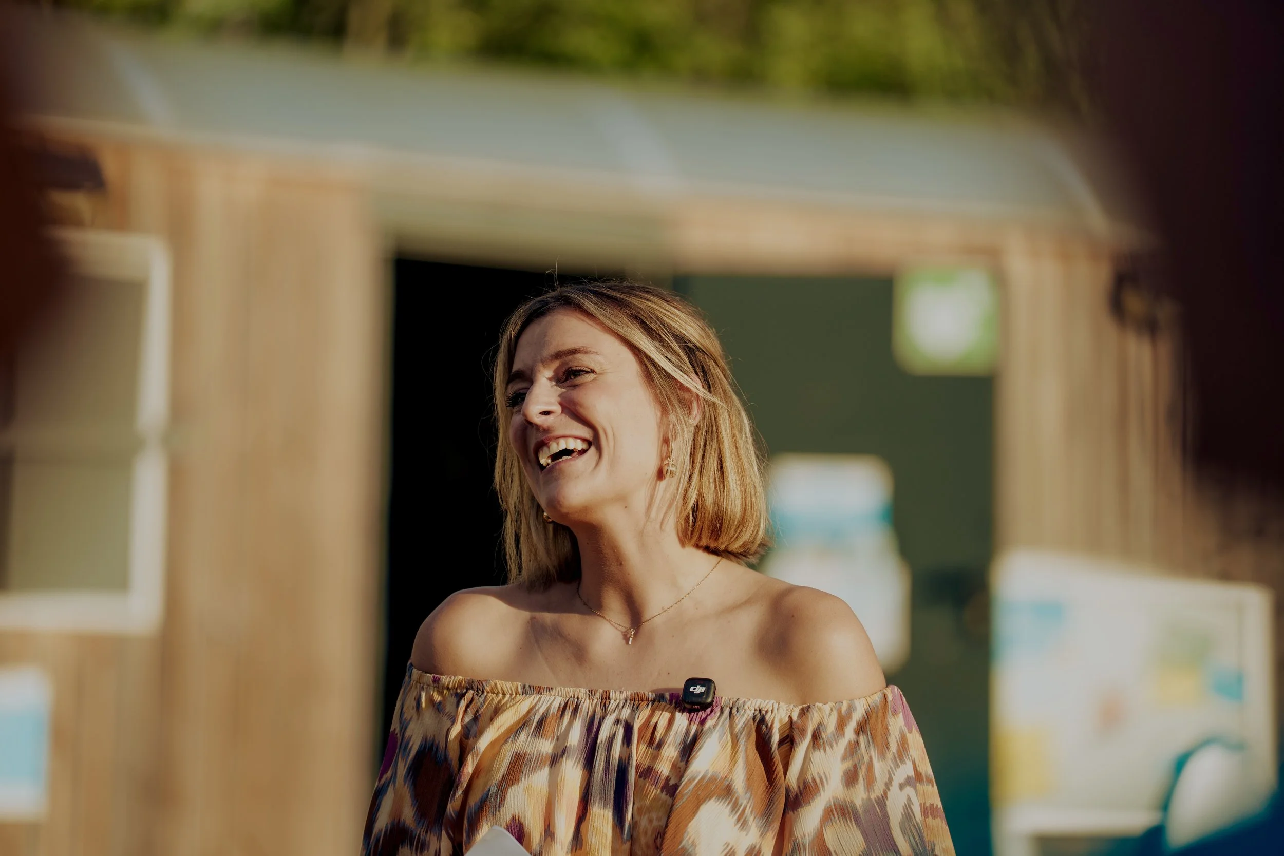 Eine lachende Frau mit Schulterlänge dunkelblonden Haaren im Freien, trägt ein bunt gemustertes Off-Shoulder-Kleid und eine Halskette.