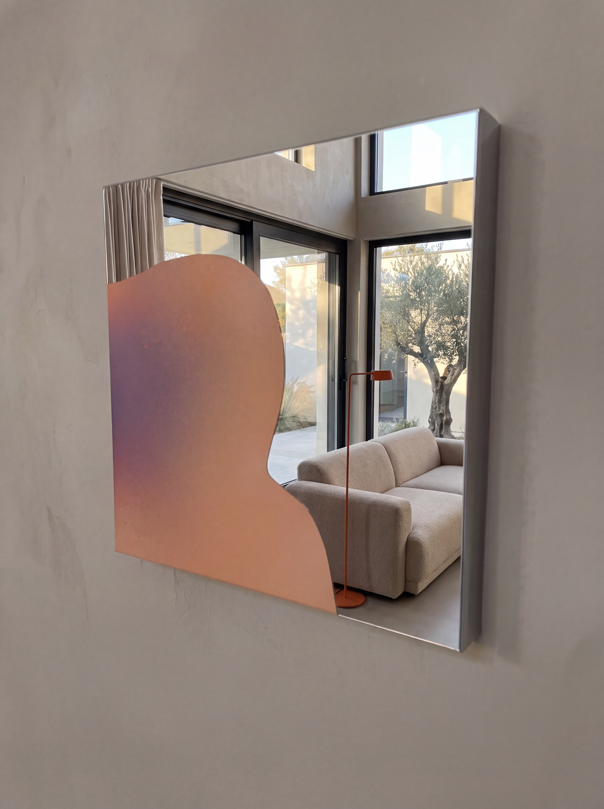 Modernes Spiegel Artwork, das in sanften Pastellfarben auf spiegelpoliertem Edelstahl gestaltet ist.  Man sieht im Spiegel ein Loft Apartment mit Blick auf Olivenbäume im Garten, davor ein Bouclé-Sofa, hochwertige Innenarchitektur.