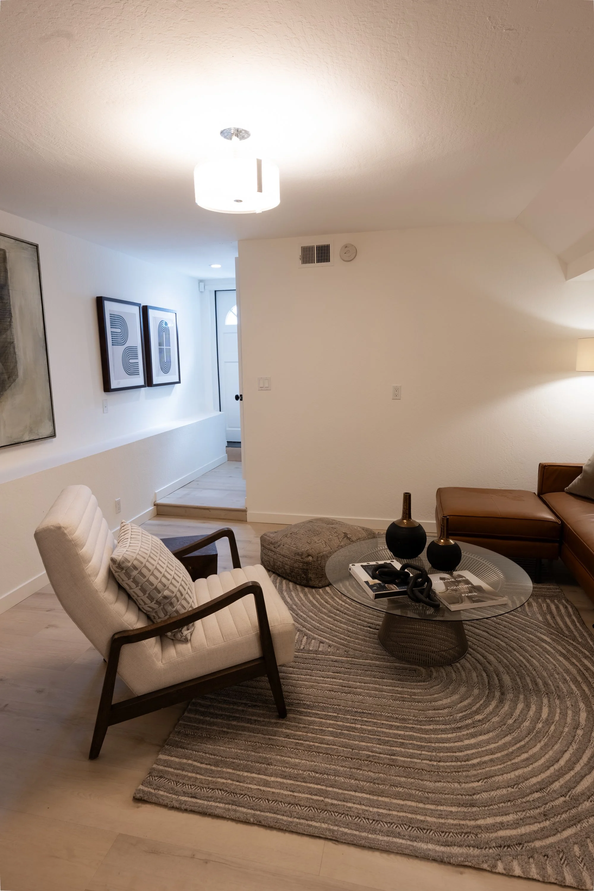 Living room with beige armchair, brown leather sofa, circular glass coffee table, area rug, decorative vases, wall art, and a ceiling light.