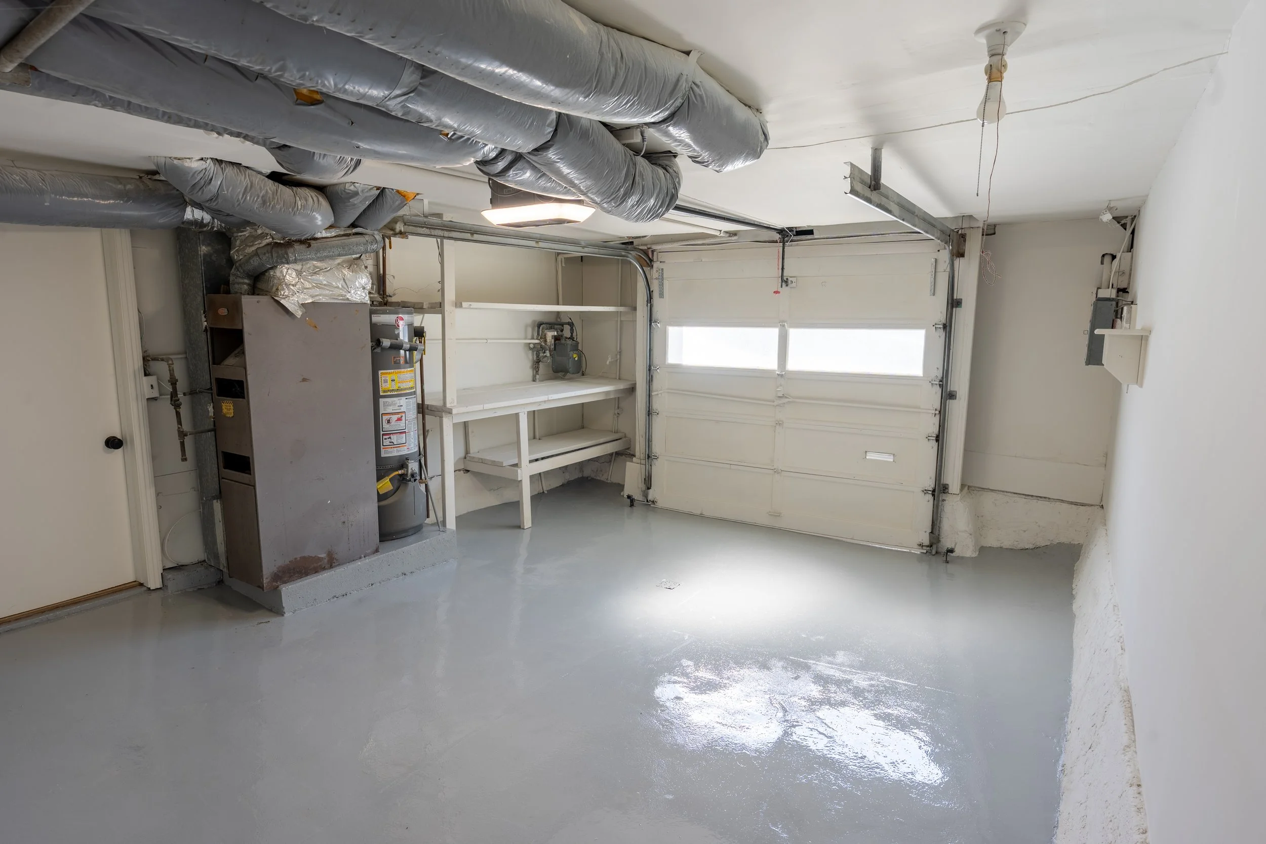 Empty garage with a concrete floor, a white garage door with windows, and exposed ductwork on the ceiling. There is a furnace and other mechanical equipment on the left side near the wall.