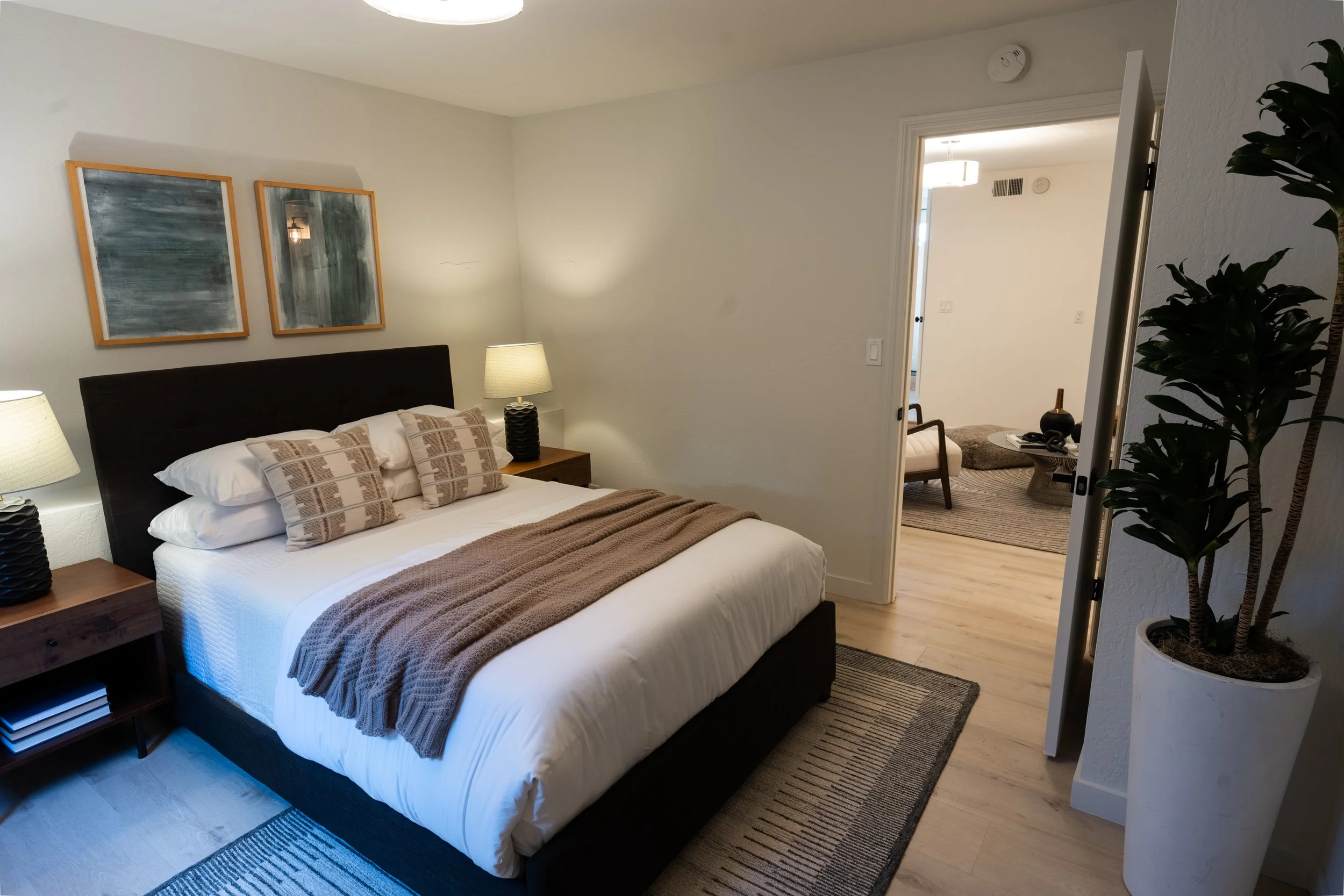 A bedroom with a bed featuring white sheets, beige pillows, and a brown blanket. There are two matching bedside lamps on wooden nightstands, and two abstract paintings hanging above the headboard. An open door leads to a seating area with a chair, a 