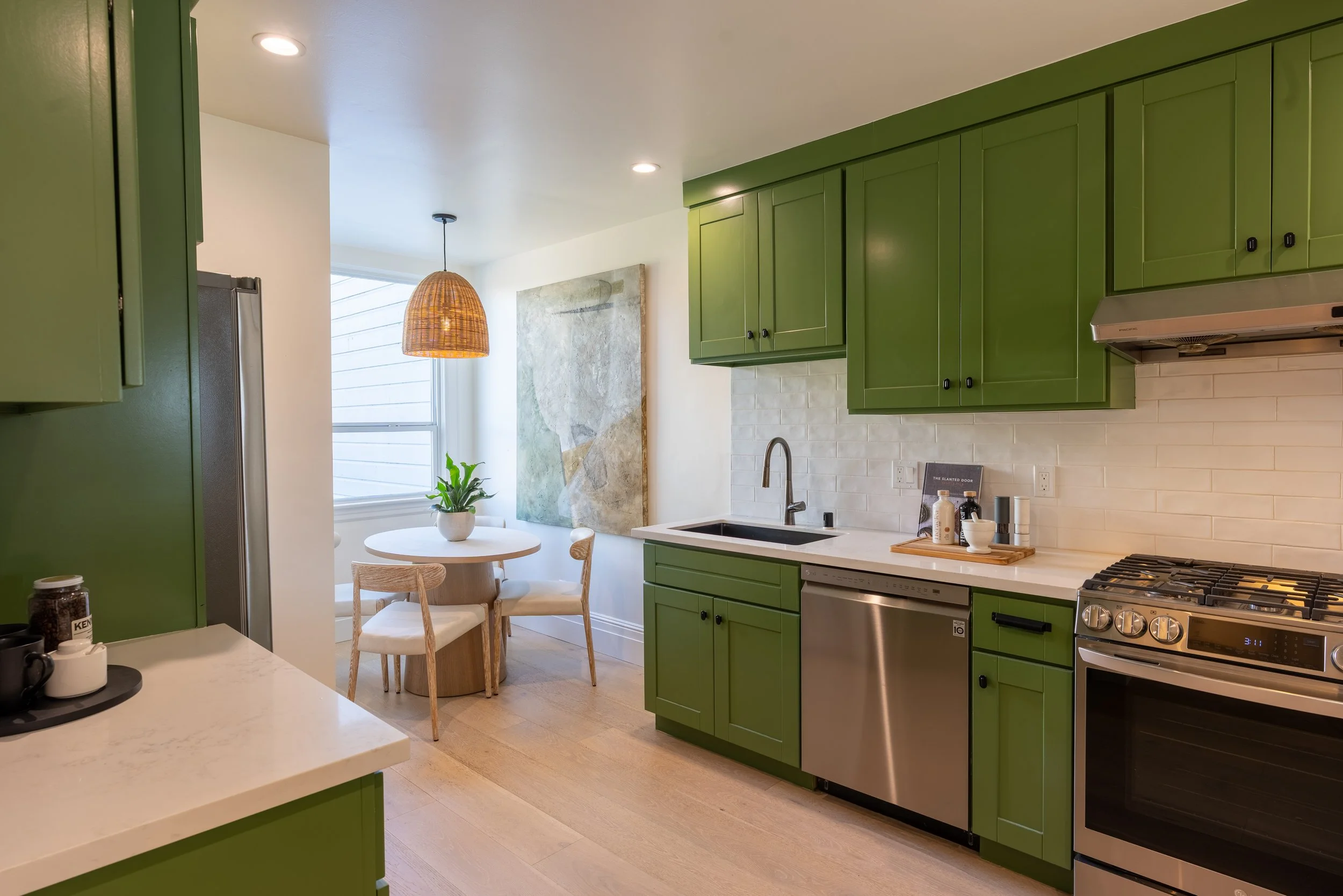 Modern kitchen with green cabinets, white countertop, stainless steel appliances, and a round dining table with chairs. A large window with blinds and a pendant light are visible in the background.