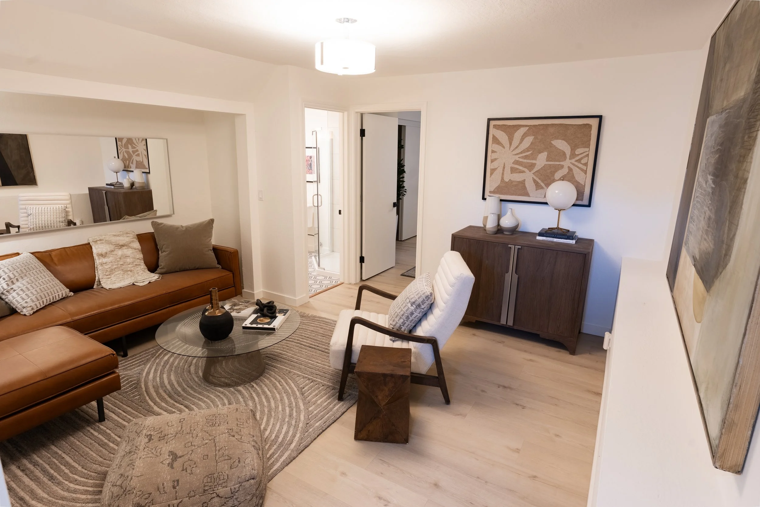Living room with a brown leather sofa, a white armchair, round glass coffee table, sideboard with decorative vases and a lamp, framed abstract artwork on the wall, mirror reflecting dining area, door to bathroom, and hardwood floors.