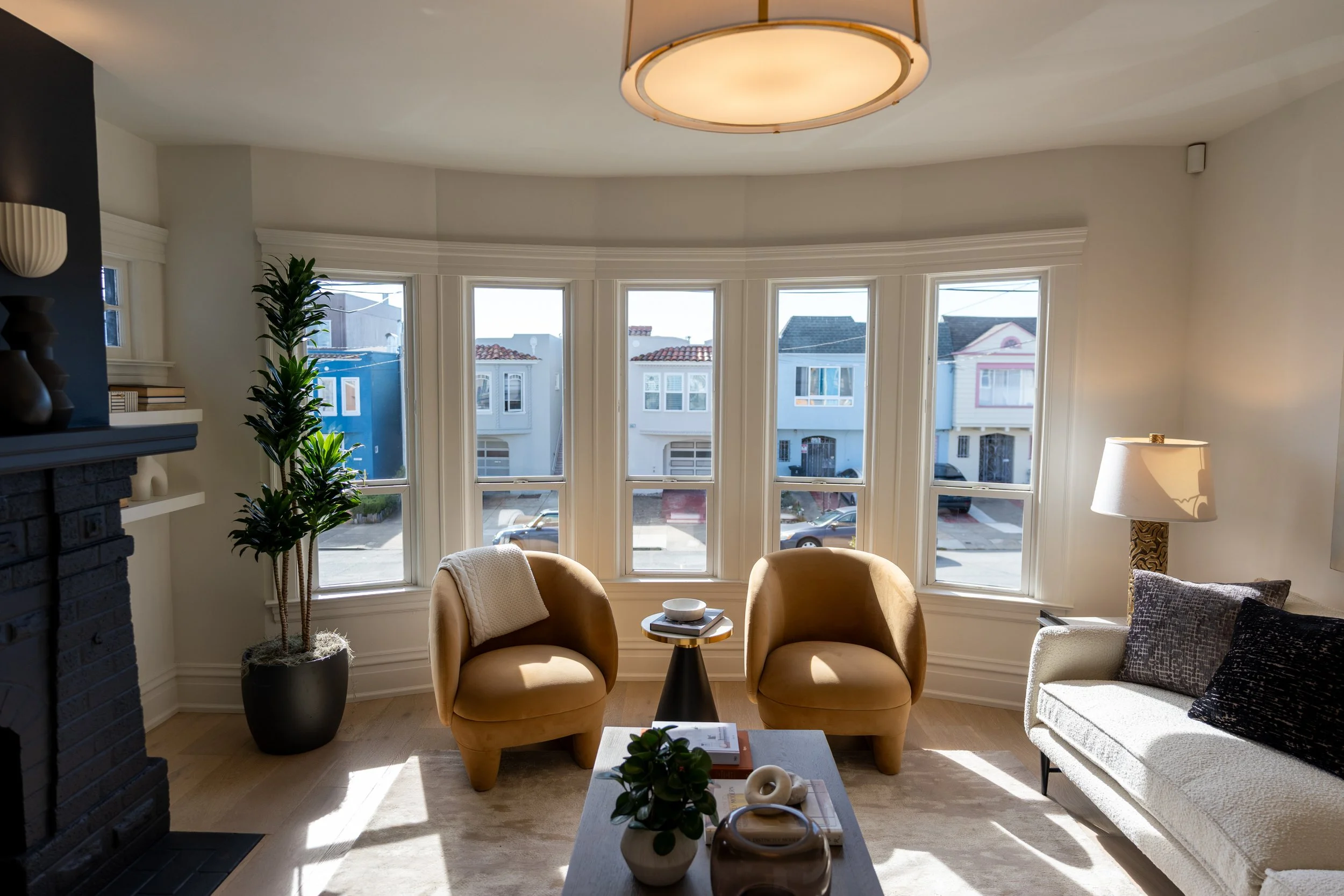 Living room with large bay window, two tan chairs, a white sofa with black pillow, potted plant, side table, and ceiling light.