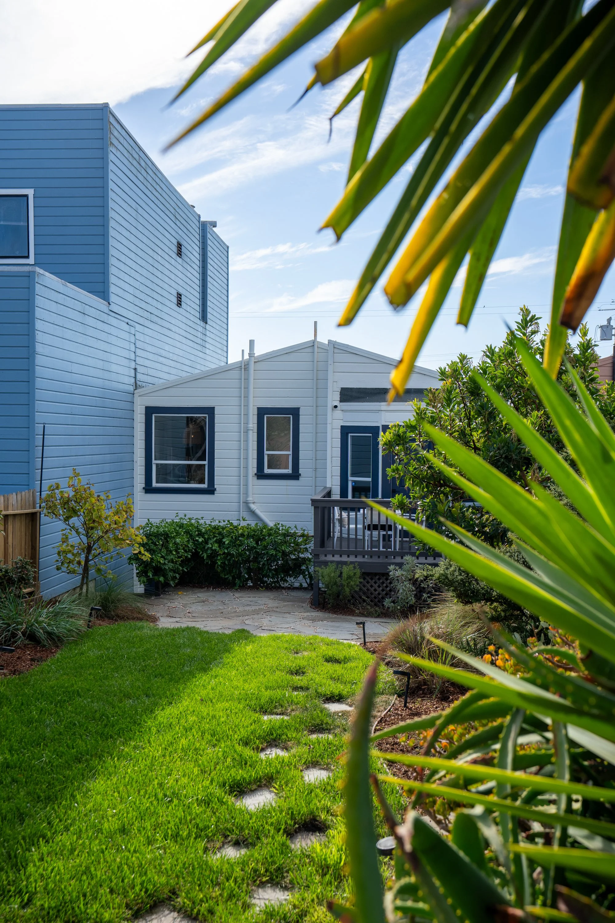 View of a backyard garden with lush green grass, various plants, and a stone pathway leading to a deck. There are two buildings in the background, one painted blue and the other white, with a partly cloudy sky overhead. Large green palm-like leaves a