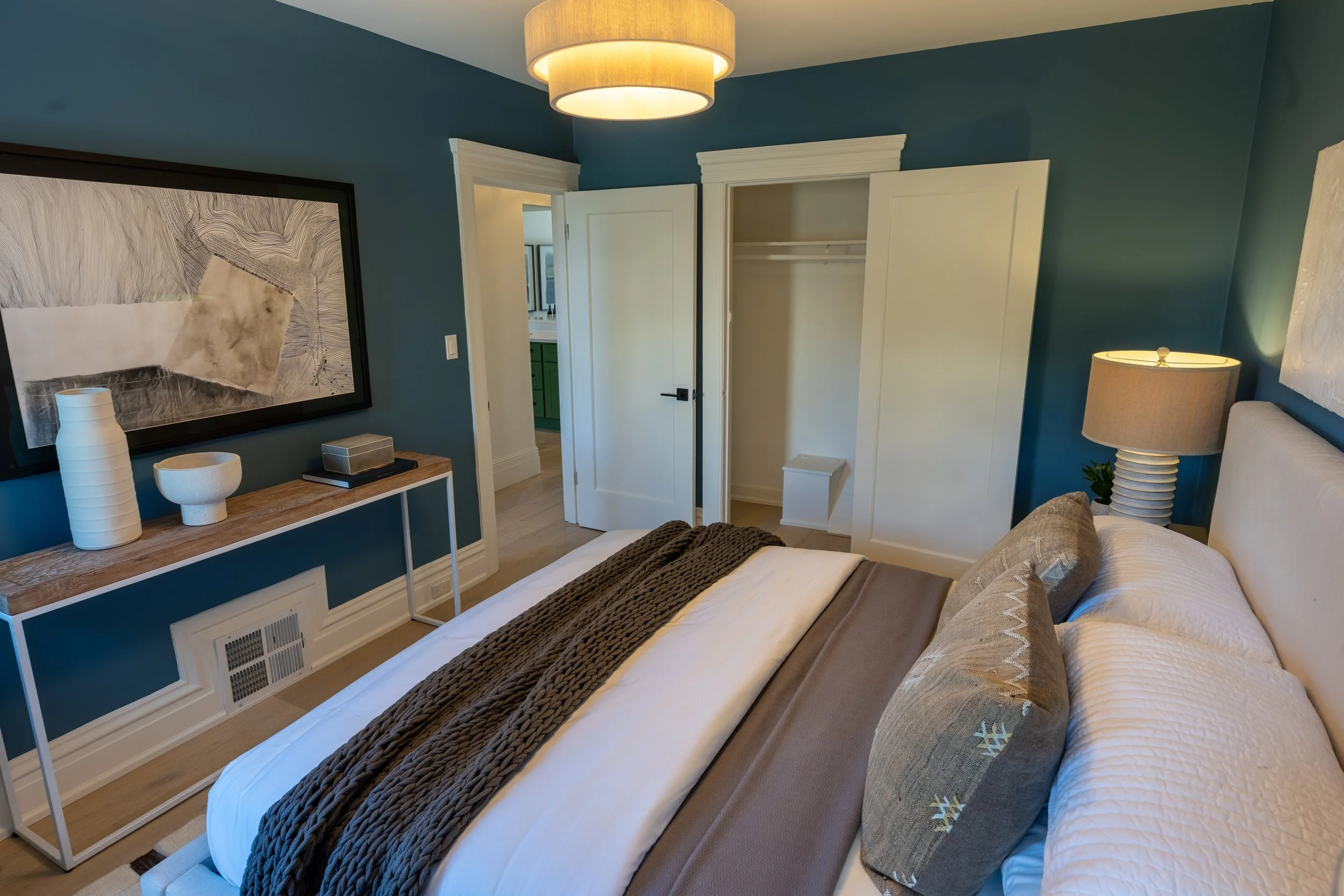 A cozy bedroom with a white bed, gray and patterned pillows, brown blanket, and a nightstand with decorative vases. The walls are painted teal, with a large black and white abstract artwork. There's a small open closet with a white door, a lamp on th