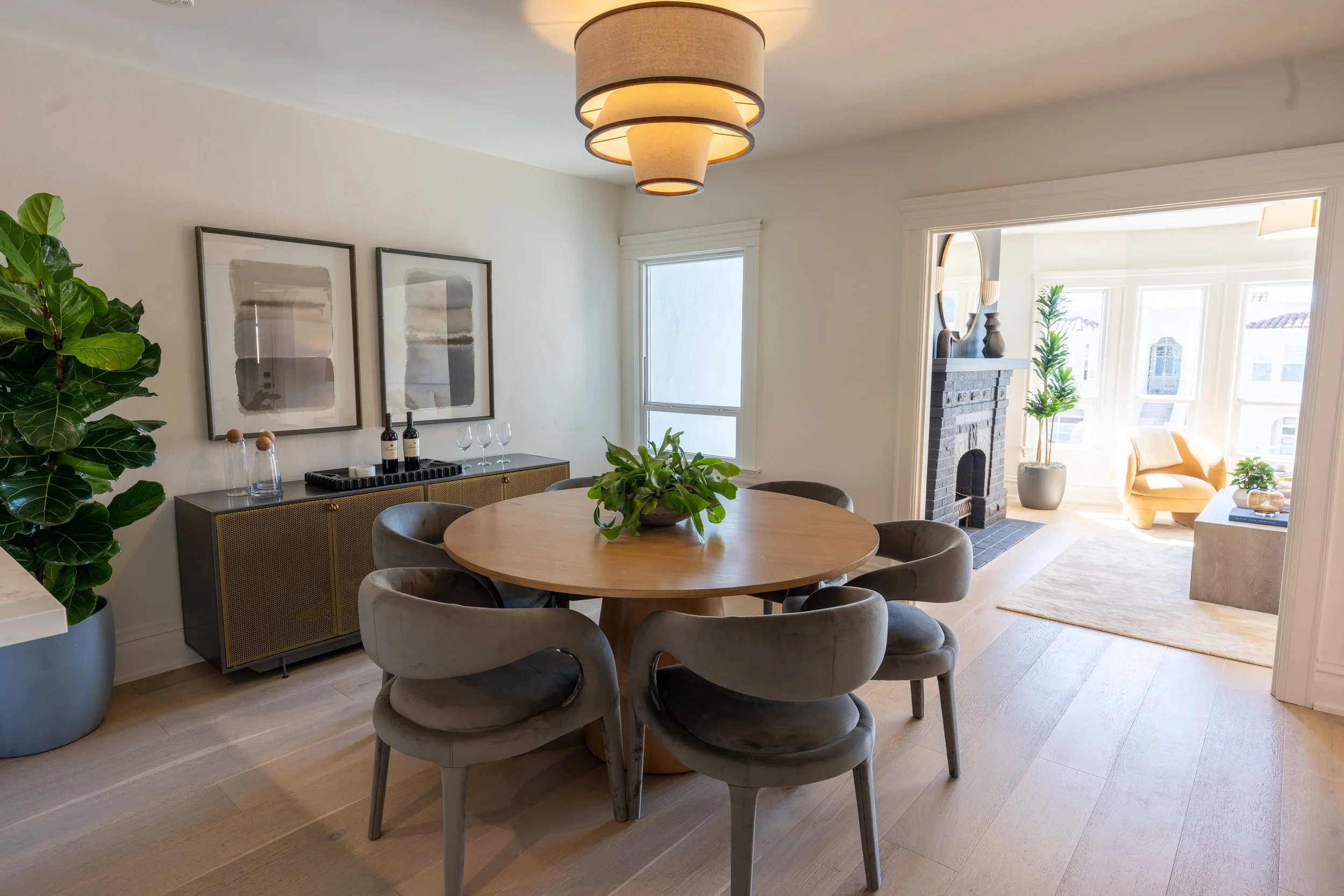A dining room with a round wooden table surrounded by six gray upholstered chairs. There is a large potted plant on the left, two abstract art pieces on the wall, a credenza with wine bottles and glasses, and a hanging light fixture. A bright sunlit 