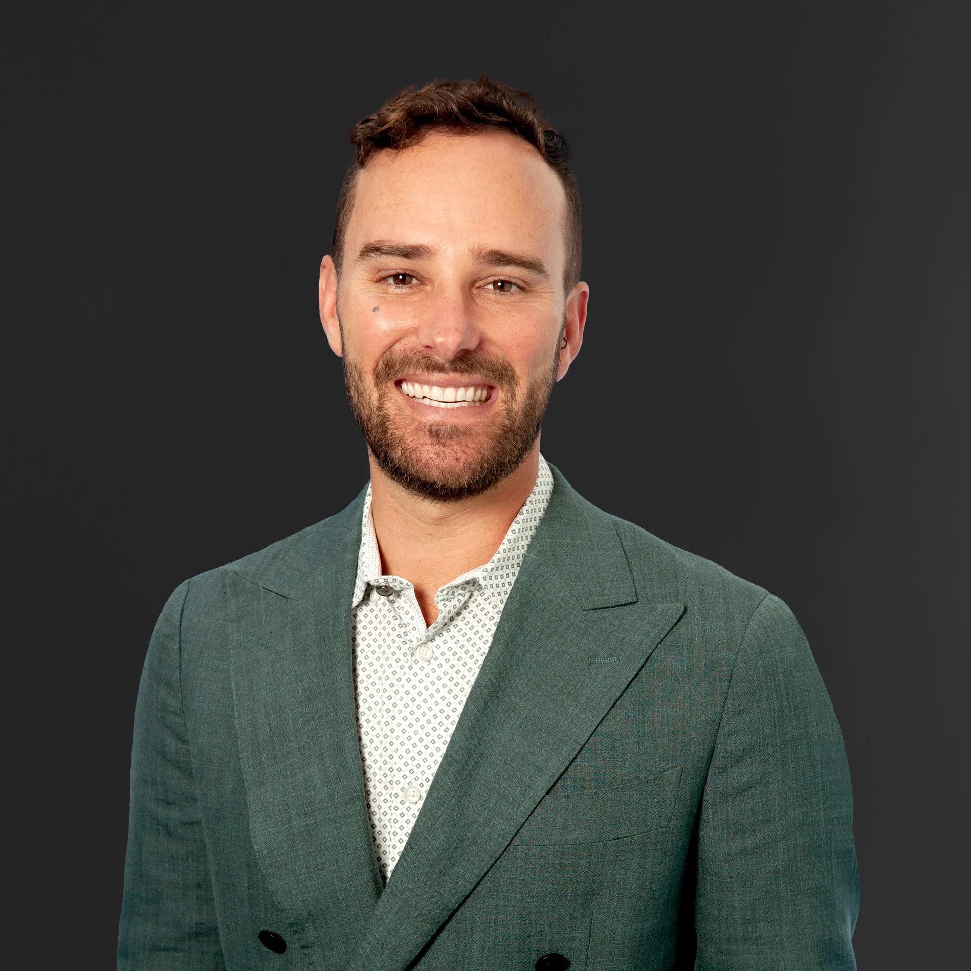Close-up of a smiling man with a beard, wearing a green blazer and white patterned shirt, against a dark gray background.