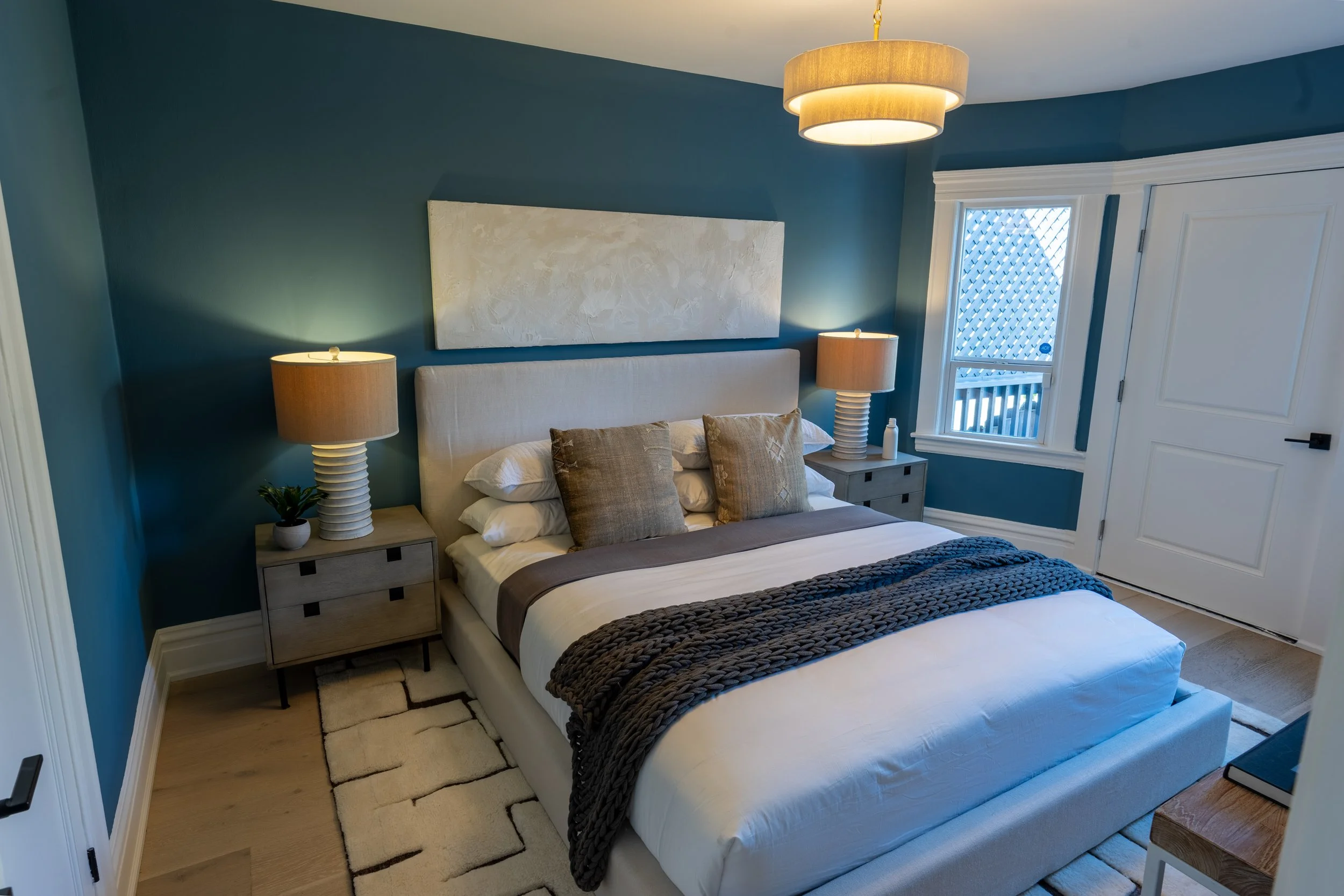 A neatly made bed with beige and brown pillows, a woven dark brown blanket, and white sheets, flanked by matching wooden nightstands with lamps, in a room with teal walls, a window with white trim, and a white door.