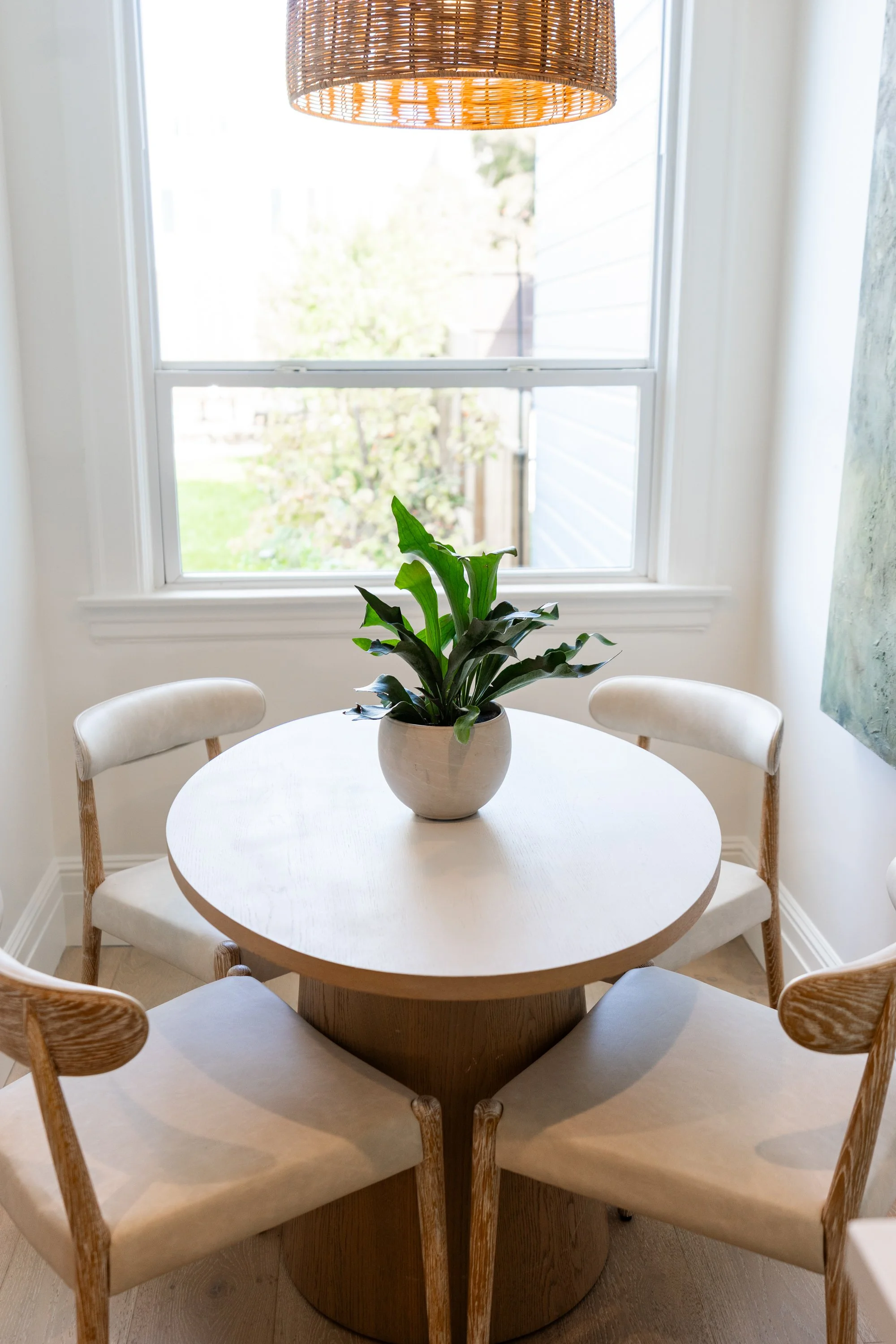 A round white table with a potted plant in the center, surrounded by five white chairs with light wood legs. In the background, a window with bright natural light, a woven pendant lamp hanging above, and a partial view of the outdoors.