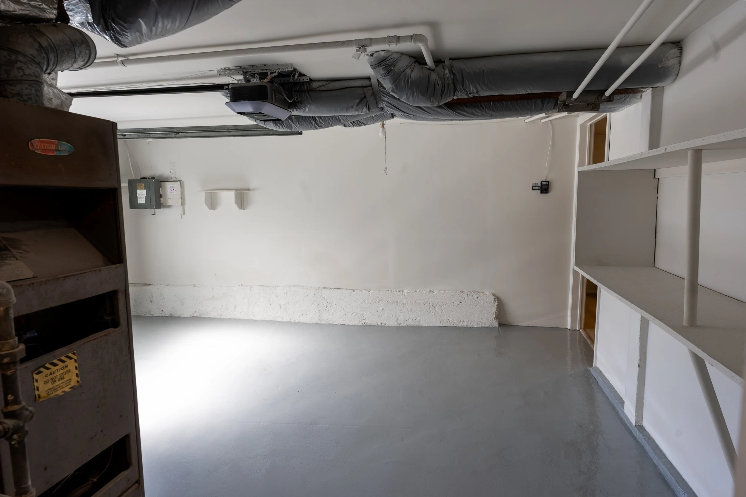 Empty laundry room or utility room with white walls, exposed ducts and pipes on the ceiling, built-in shelves on the right, and electrical panels on the back wall.