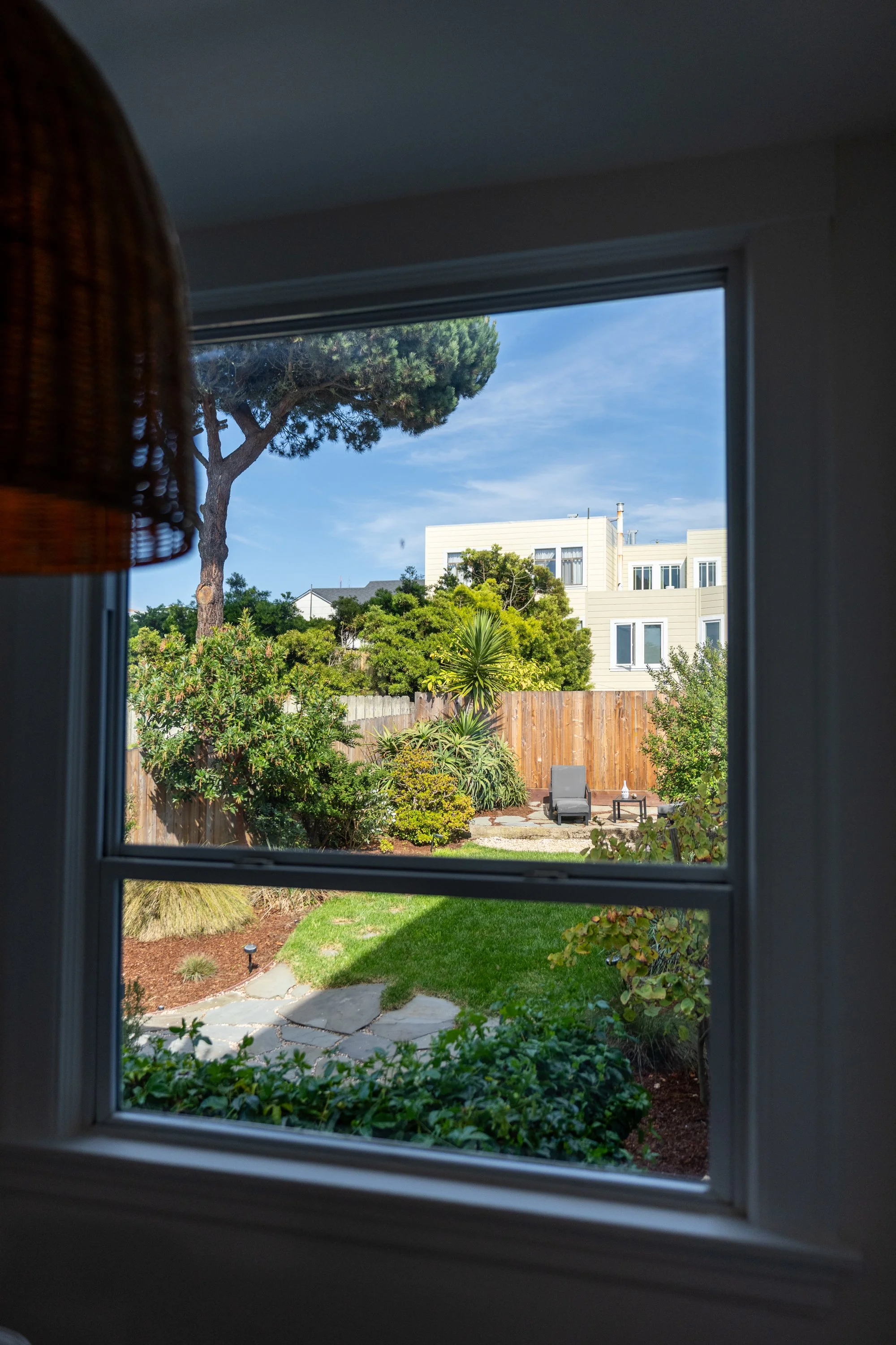 View of a backyard garden with various green shrubs and trees, a stone pathway, a patio with outdoor furniture, and a multi-story house in the background, seen through a window.