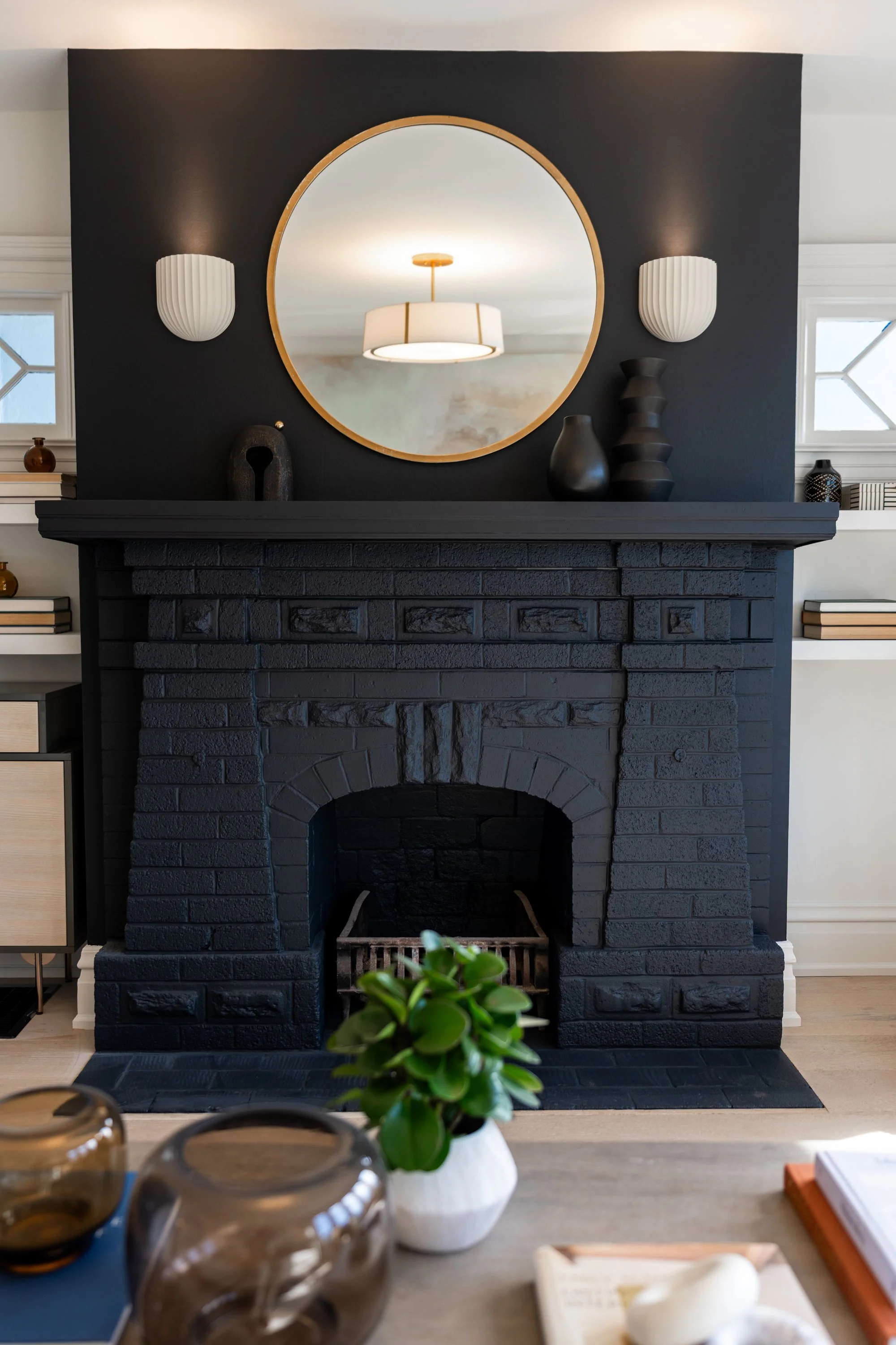 Black brick fireplace with a large round mirror above it, flanked by white wall sconces, with decorative vases and objects on the mantle and shelves, and a table with plants and objects in the foreground.