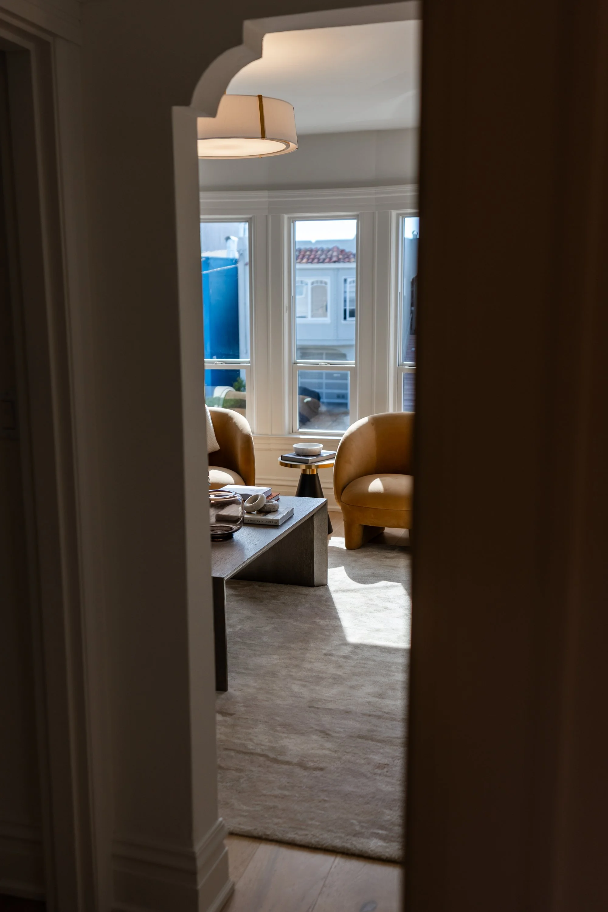 Interior view of a living room, seen through a doorway, with two yellow armchairs, a small round table, a stack of books, a bowl, and a window with a view of neighboring houses and a blue fence.